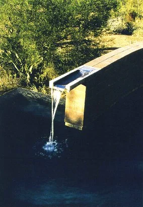 Modern outdoor fountain with water flowing into a pool, surrounded by vegetation.