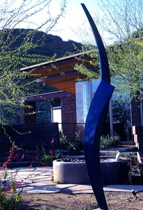 A modern outdoor sculpture in front of a building, surrounded by desert landscaping and mountains in the background.