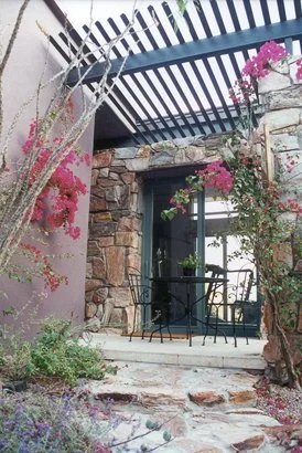 Stone patio with metal table and chairs, surrounded by flowering plants and a pergola overhead.
