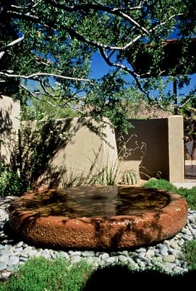 Garden with round stone water feature surrounded by pebbles, plants, and trees in a landscaped area.