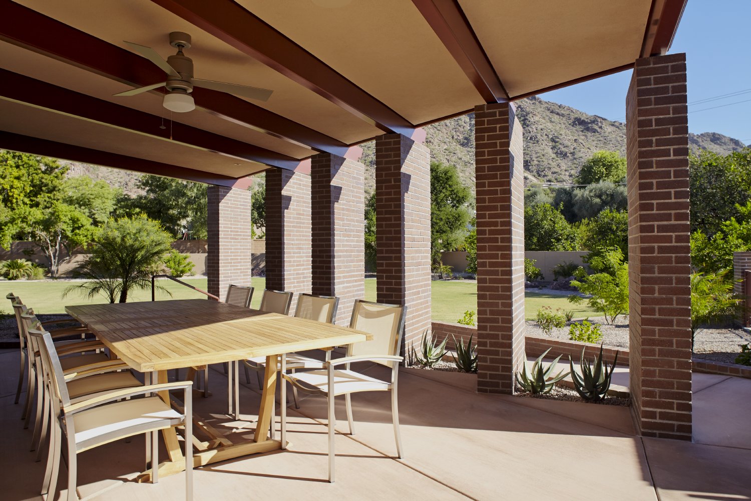 Covered patio for an outdoor dining experience featuring, brick columns, ceiling fan, and view of a garden with trees and a mountain backdrop.