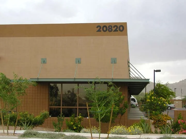 Modern beige office building, surrounded by greenery and trees, under a cloudy sky.