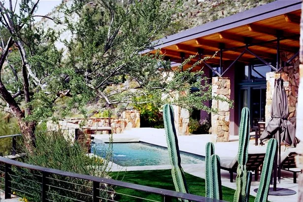 A modern desert home with stone accents and the roof extending over the pool, featuring a backyard pool surrounded by cacti and desert landscaping.