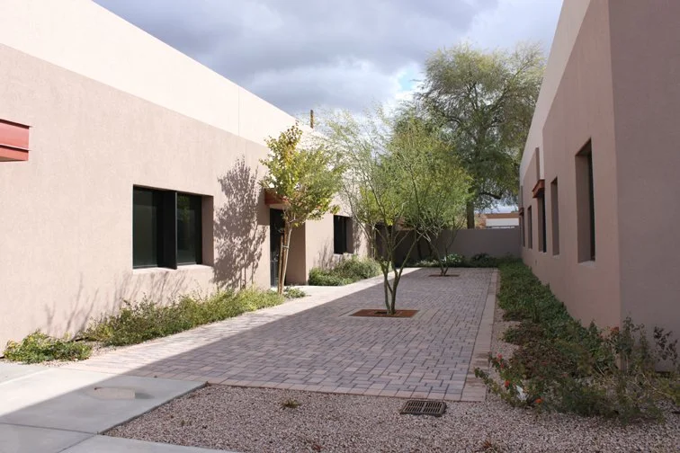 Outdoor courtyard between two stucco buildings with brick walkway, small trees, and shrubs.