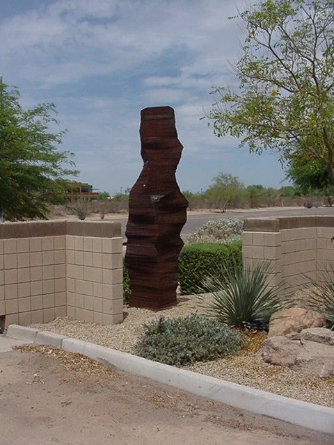 Outdoor metal sculpture with jagged edges on a landscaped stone area surrounded by low beige walls and desert plants.