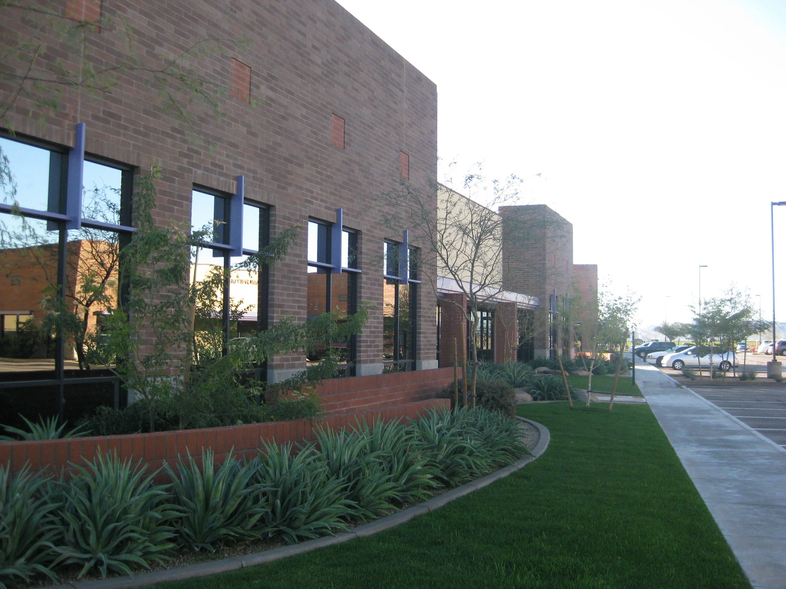 Side view of a modern brick commercial office building with large windows, landscaped garden beds, green grass, and a parking lot with vehicles in the background.