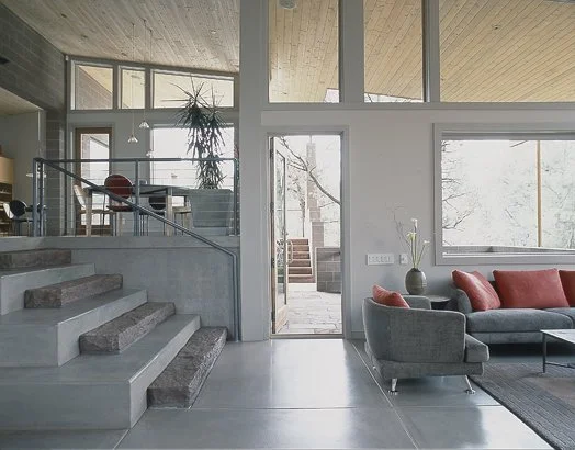 Modern living room with gray sectional sofa, red cushions, concrete steps, large windows, and wooden ceiling, featuring minimalistic interior design.
