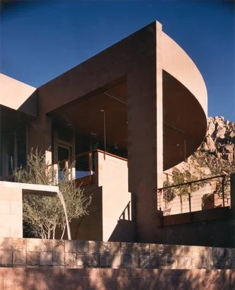 Modern architectural building with angular and curved elements, exterior view with stone facade and desert landscaping, set against a clear blue sky.