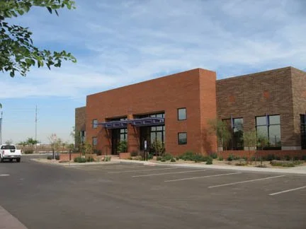 Modern commercial office building with brick façade, large windows, and empty parking lot in front, set against a clear sky.