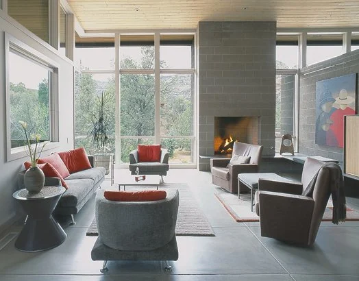Modern living room with large windows, gray sofas, red and orange cushions, armchairs, a fireplace, artwork, and decorative plants.