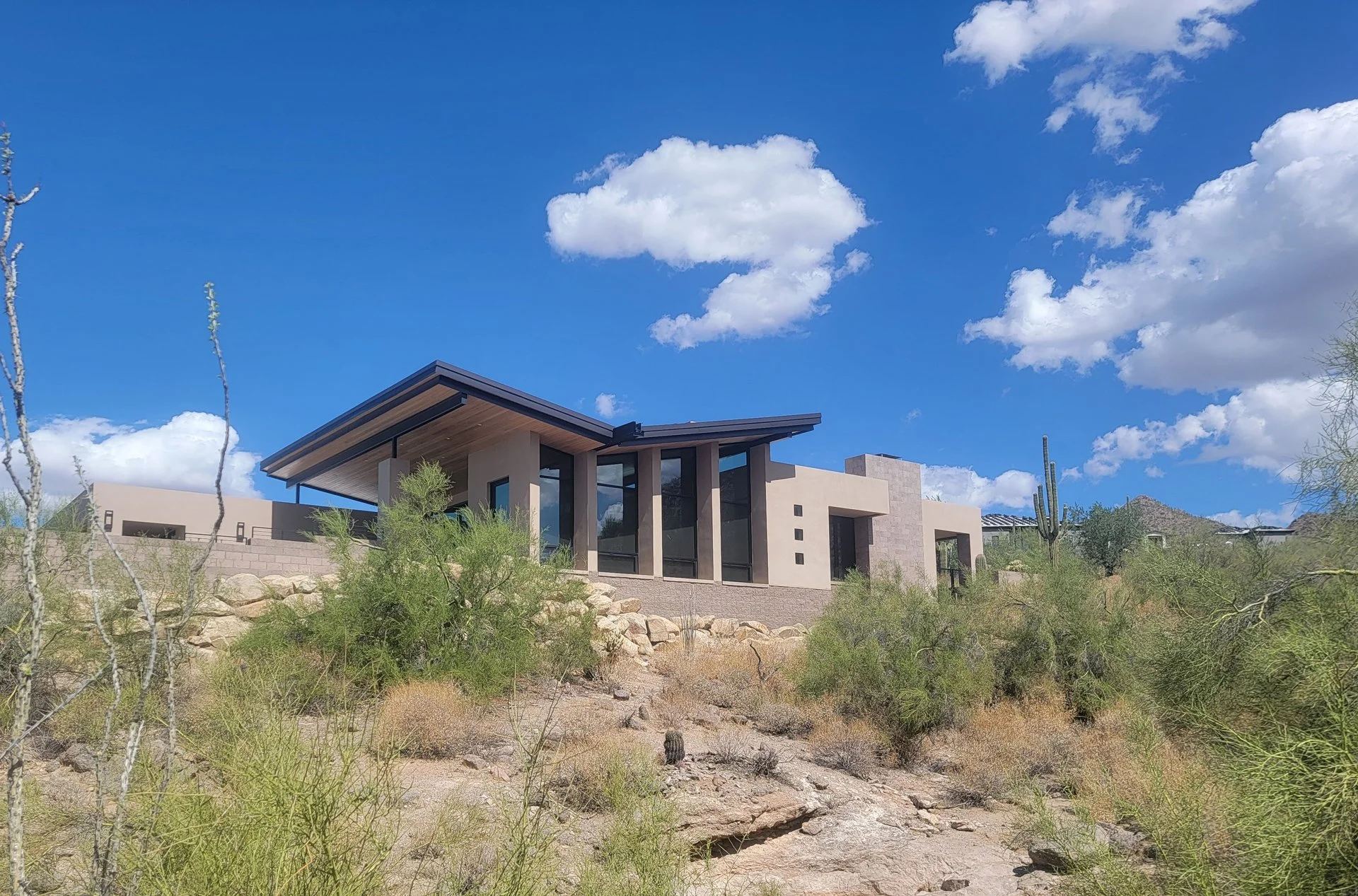 Modern house with large windows in desert landscape, surrounded by cacti and shrubs under a blue sky with clouds.