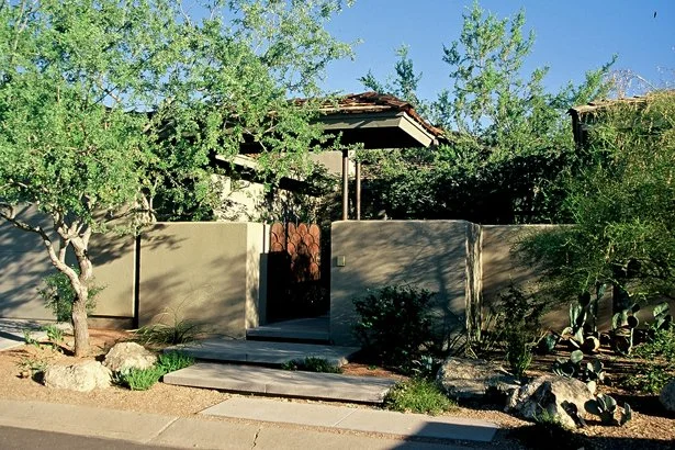 Contemporary house with a wooden gate and stucco walls surrounded by desert landscaping with rocks, cactus, and trees.