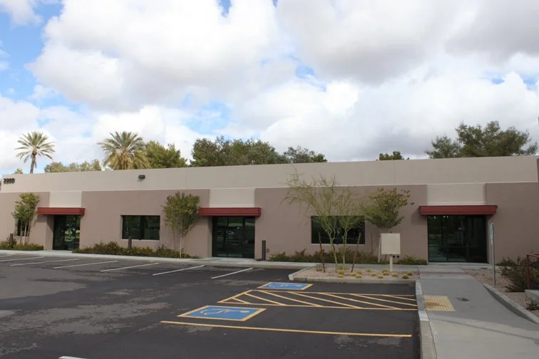 Single-story office building with red awnings, surrounded by a parking lot and trees; the sky is partly cloudy.