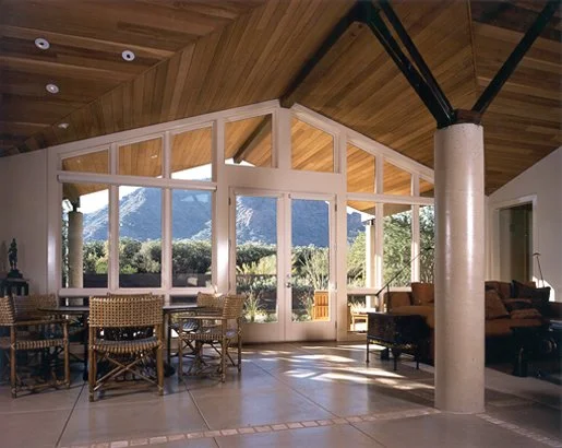 Interior of a spacious room with large windows showcasing a scenic mountain view. The room features a wooden ceiling, a dining area with wicker chairs, and a cozy seating area to the right. Natural light floods the space, highlighting the open design