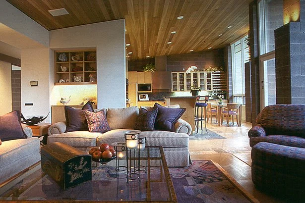 Contemporary living room with cozy furniture, wooden ceiling, and open kitchen and dining area in the background.