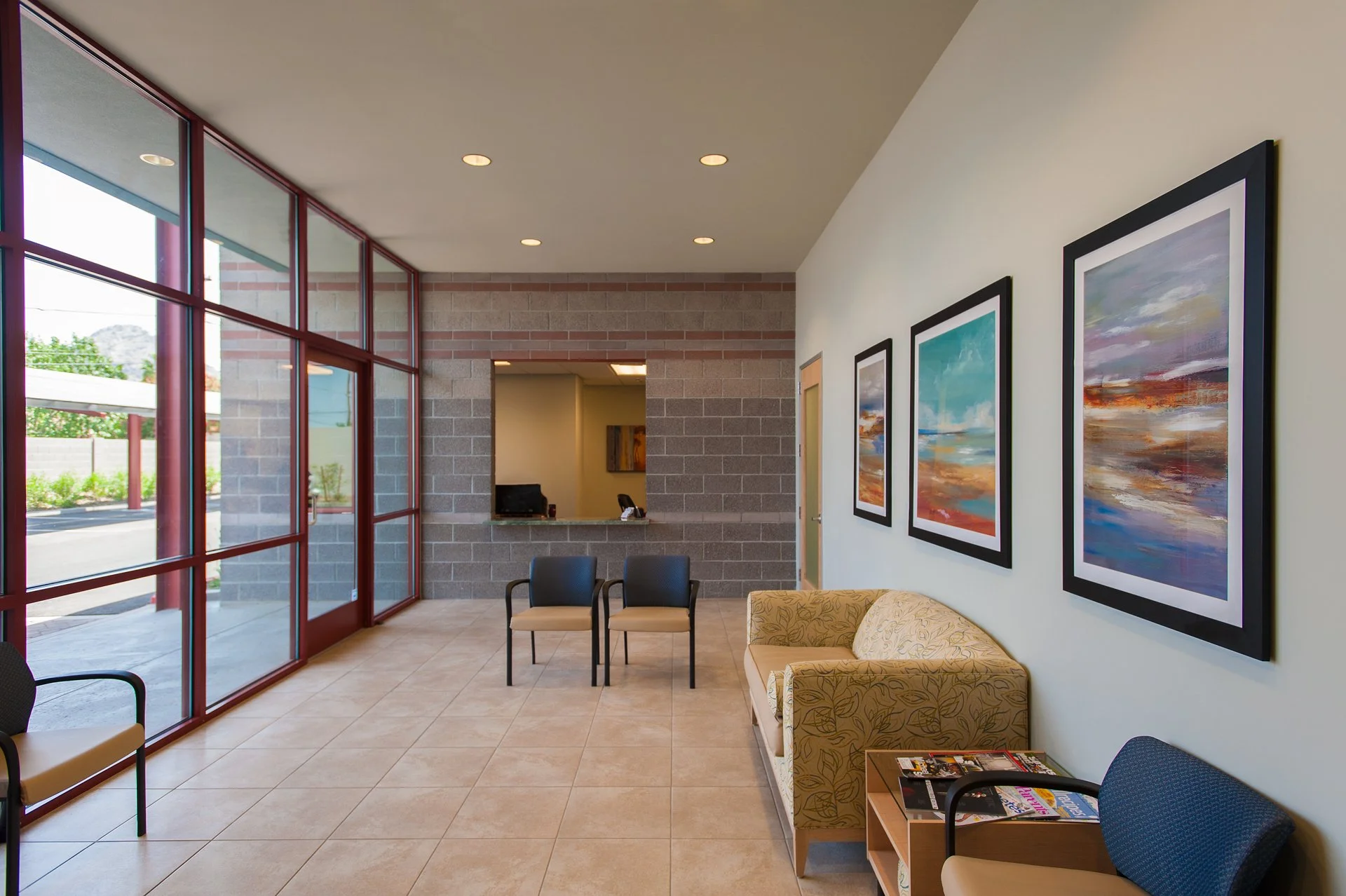 Modern medical office waiting room with large windows, chairs, abstract art on the wall, and a small table with magazines.