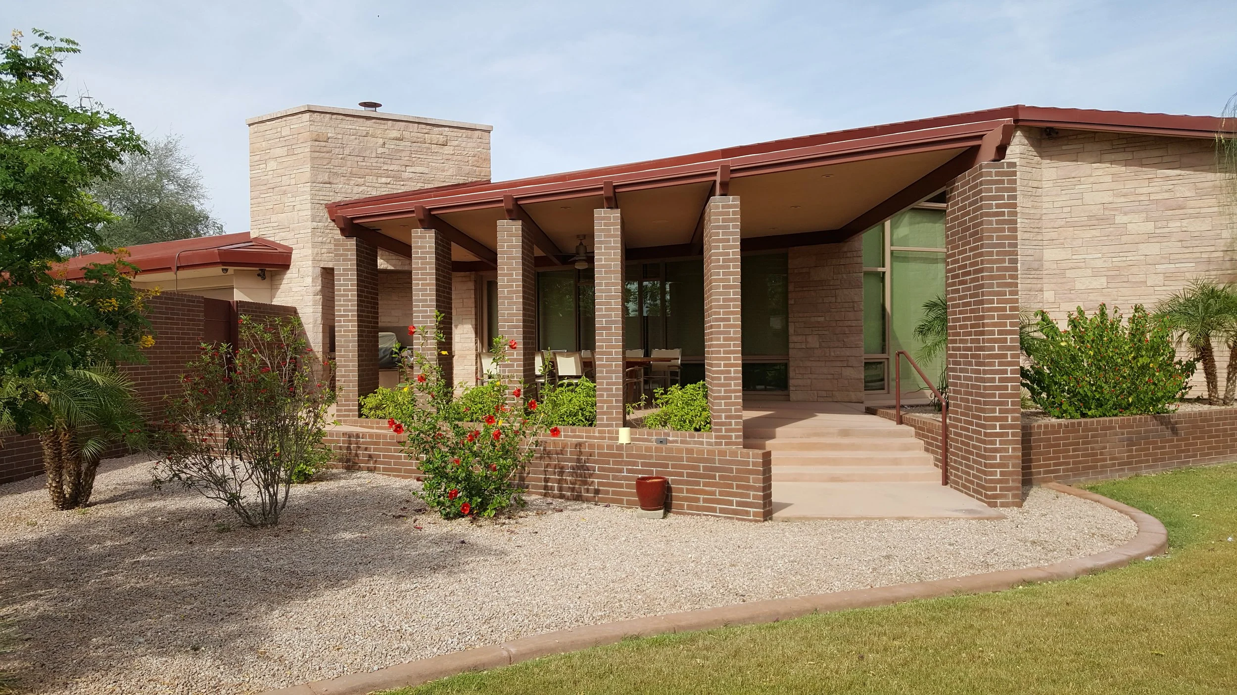 Modern stone masonry house with a covered porch held up by brick columns, surrounded by desert landscaping and shrubs, featuring red flowers.