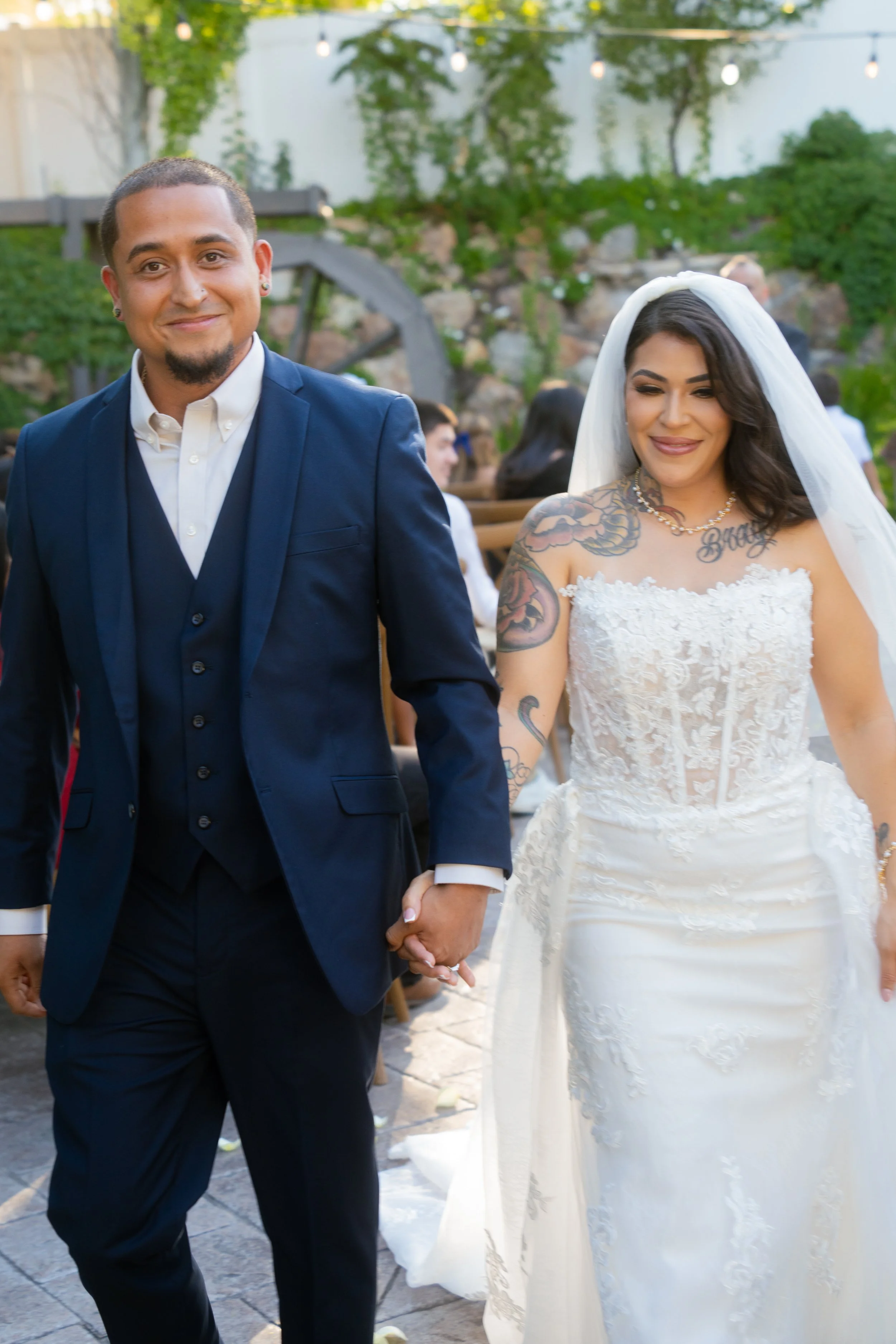 A newly married couple walking hand in hand at an outdoor wedding reception, with string lights overhead and other guests in the background.