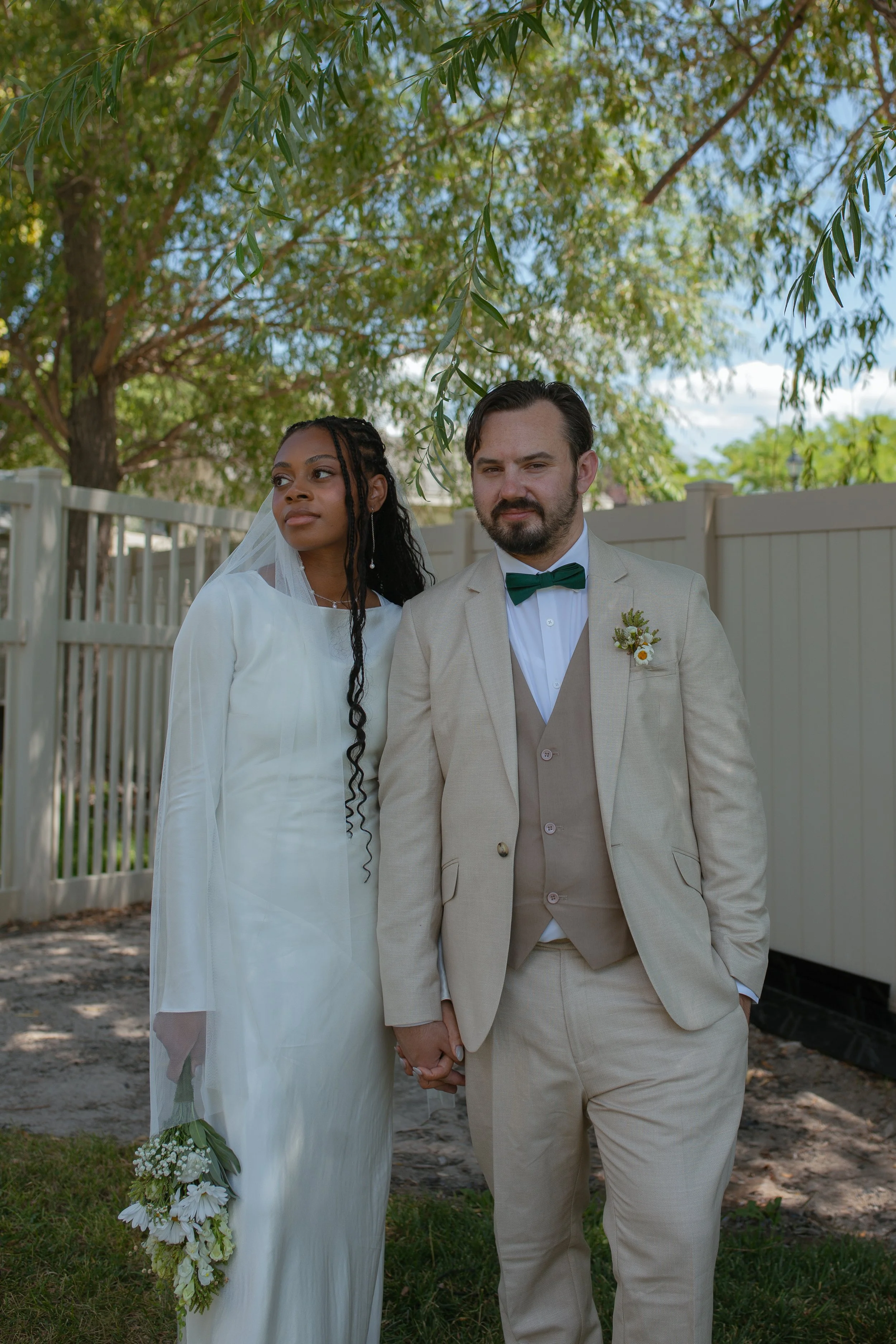 A newlywed couple standing outdoors on a sunny day, holding hands with a white picket fence and green trees in the background. The bride is wearing a white wedding dress with a veil, and the groom is wearing a beige suit with a green bow tie.