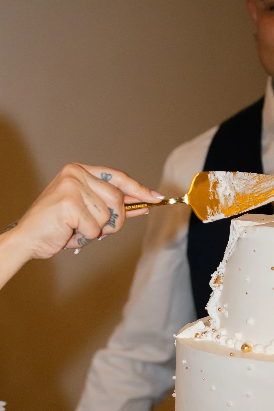 A person's hand with tattoos applying white icing to a wedding cake with gold and white pearls.