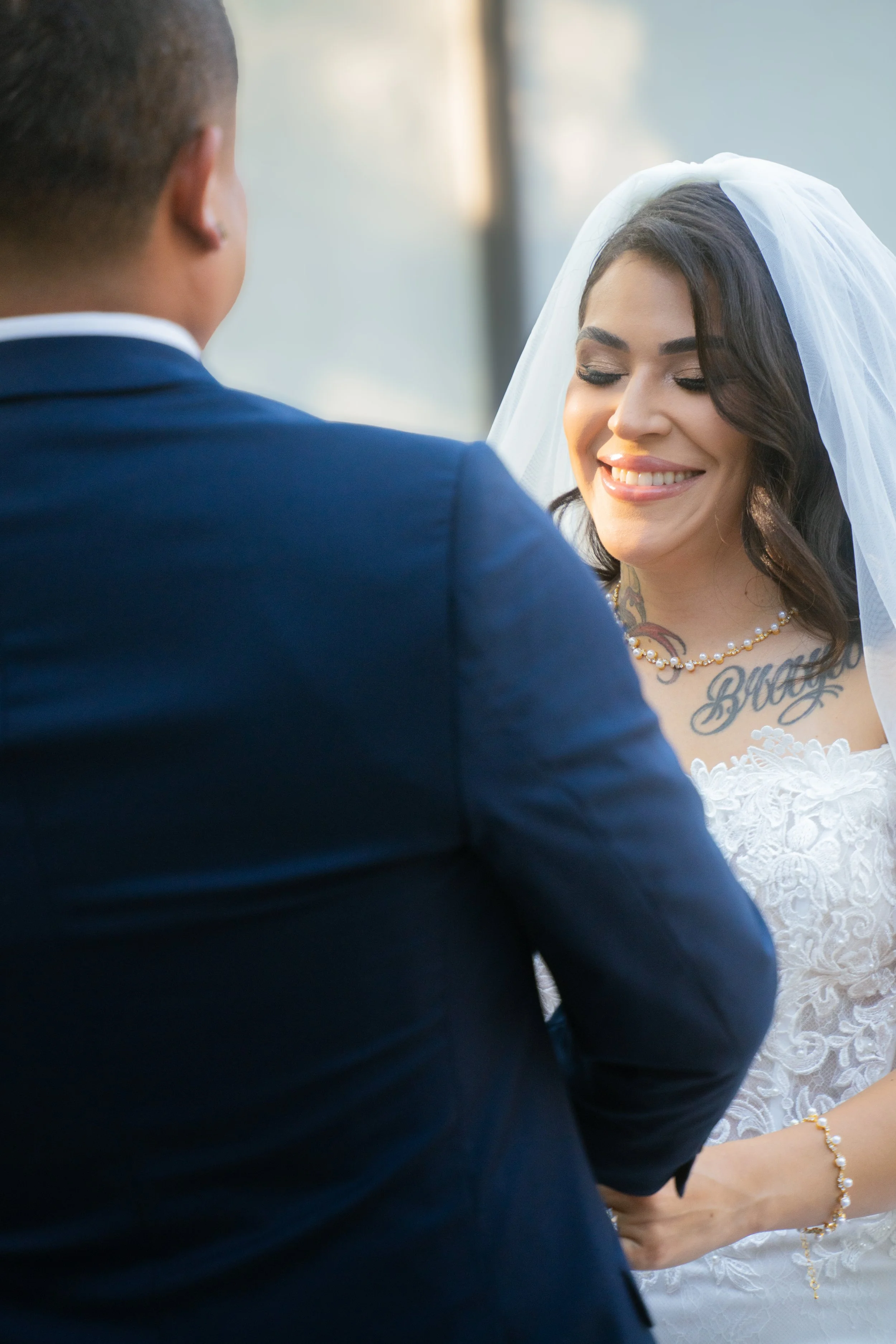 A bride and groom during their wedding ceremony, with the bride smiling and wearing a white dress, veil, and pearl jewelry, and the groom in a dark suit.