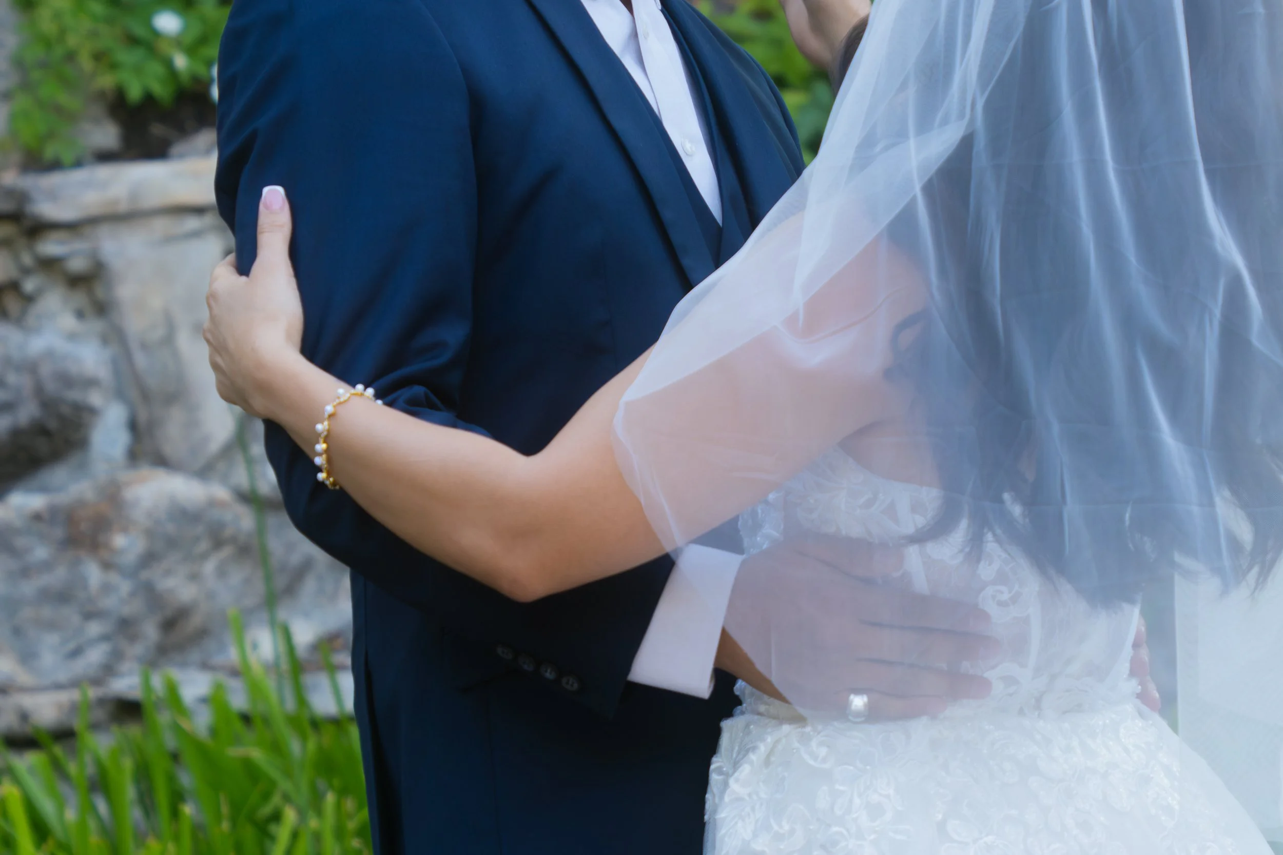 A couple sharing a dance outdoors, with the woman wearing a white dress and a tulle veil, and the man in a navy suit, surrounded by greenery.