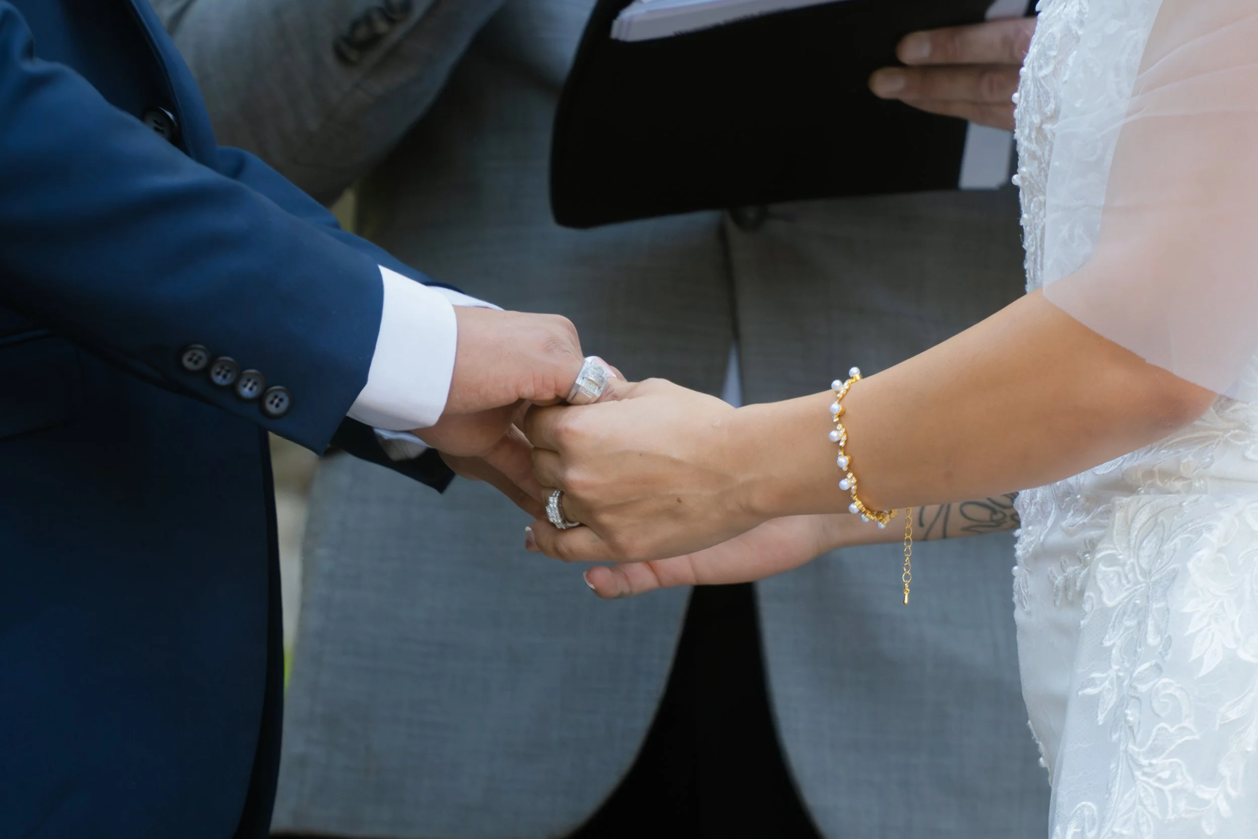 Close-up of a man and a woman holding hands during a wedding ceremony, with the man's hand wearing a wedding ring and the woman's hand adorned with jewelry. The woman is wearing a white wedding dress with lace details.
