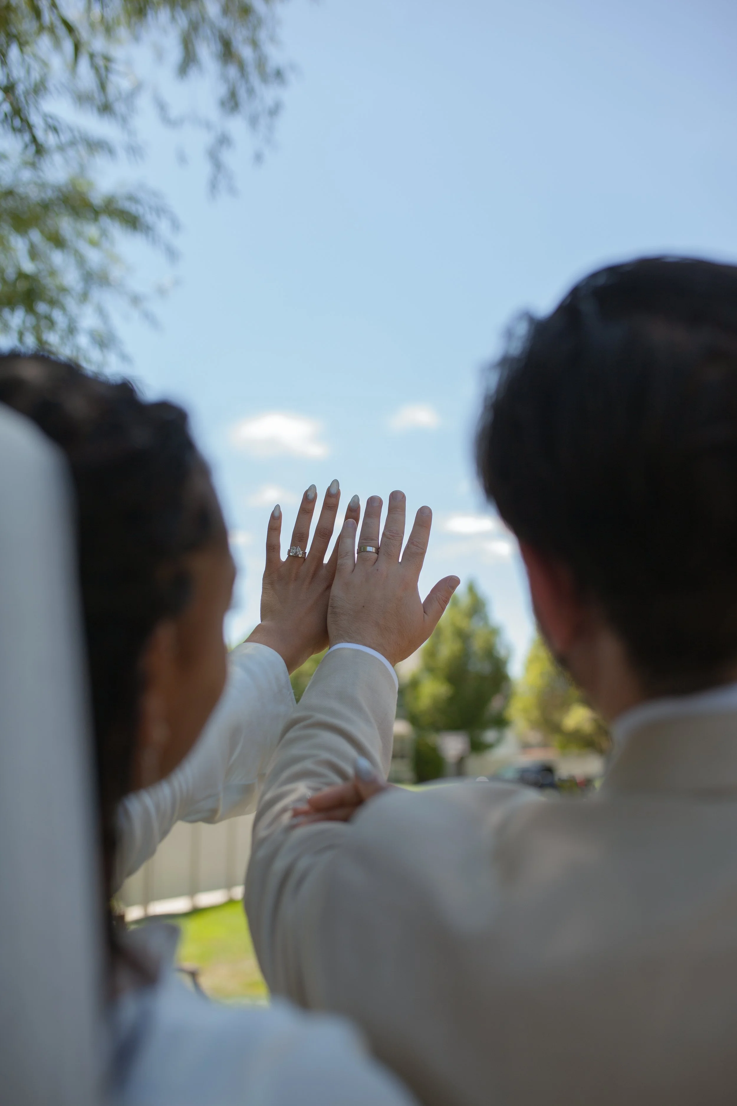 Close-up of a woman in white wedding dress with her hand raised, showing wedding rings, surrounded by two men in suits, outdoors on a sunny day.