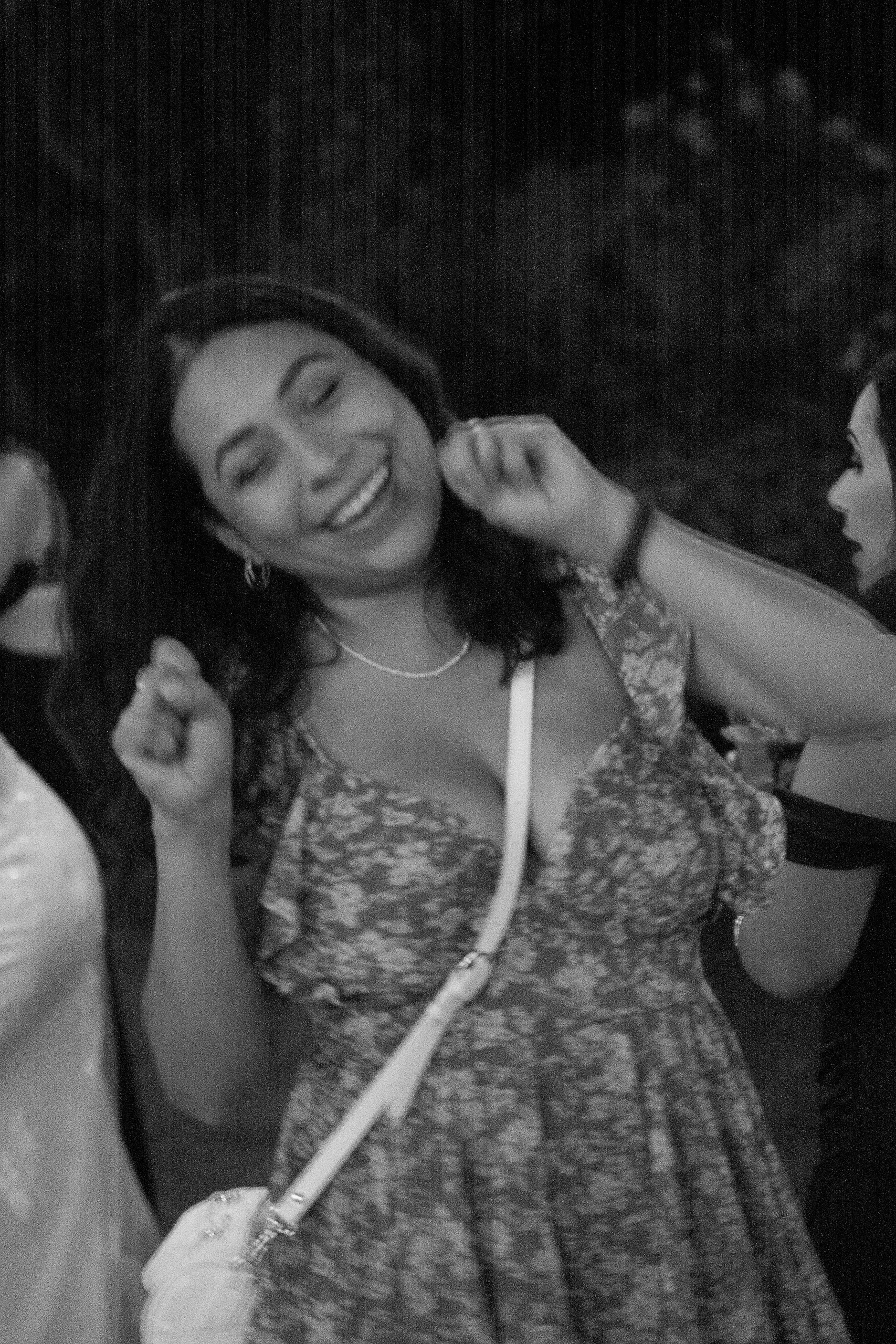A woman with long dark hair smiling and wearing earrings, with another woman partially visible on the right side of the image. The woman on the right has short hair and glasses, and they appear to be in a social setting.