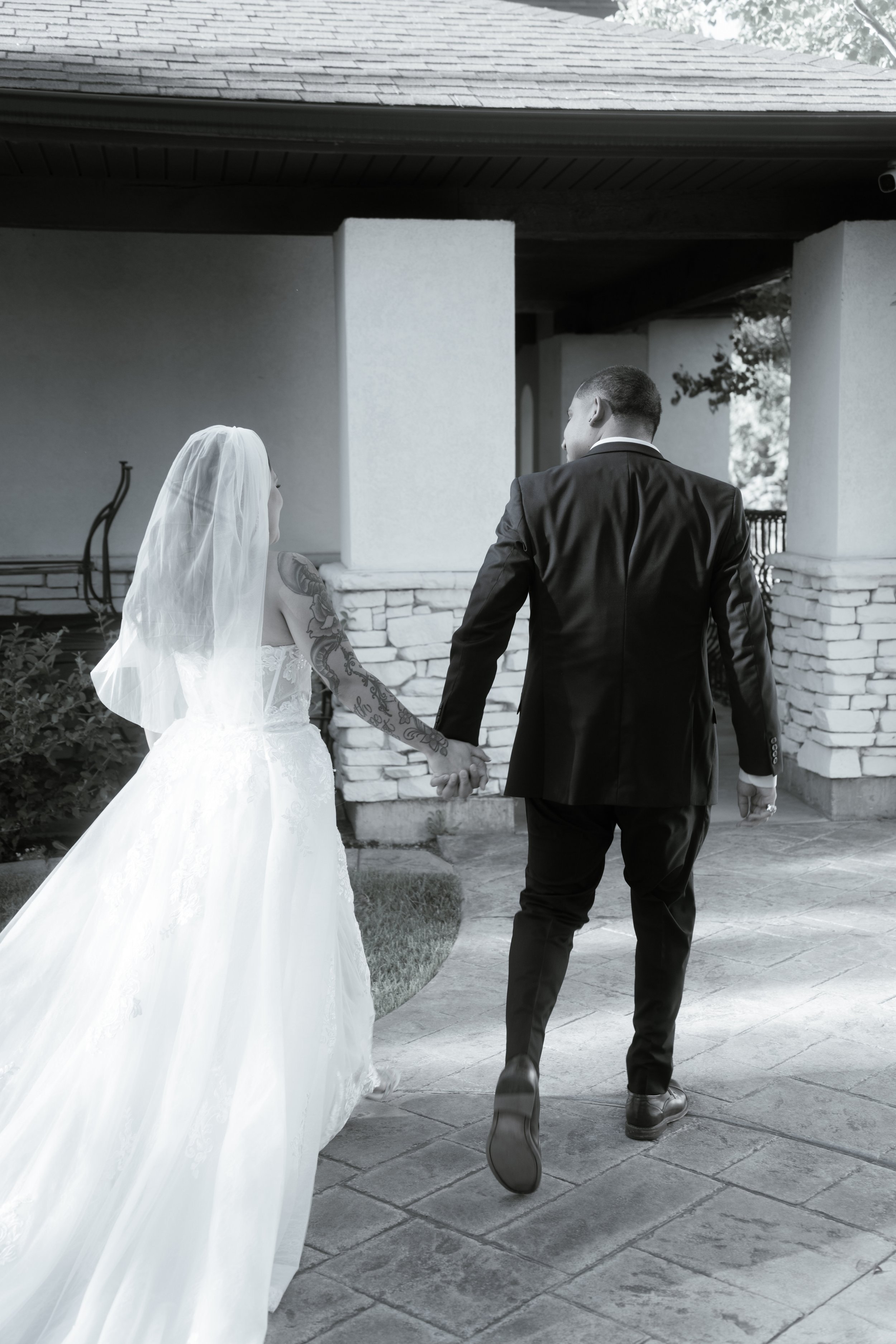 A bride and groom holding hands, walking away outside on a stone pavement with a house in the background.