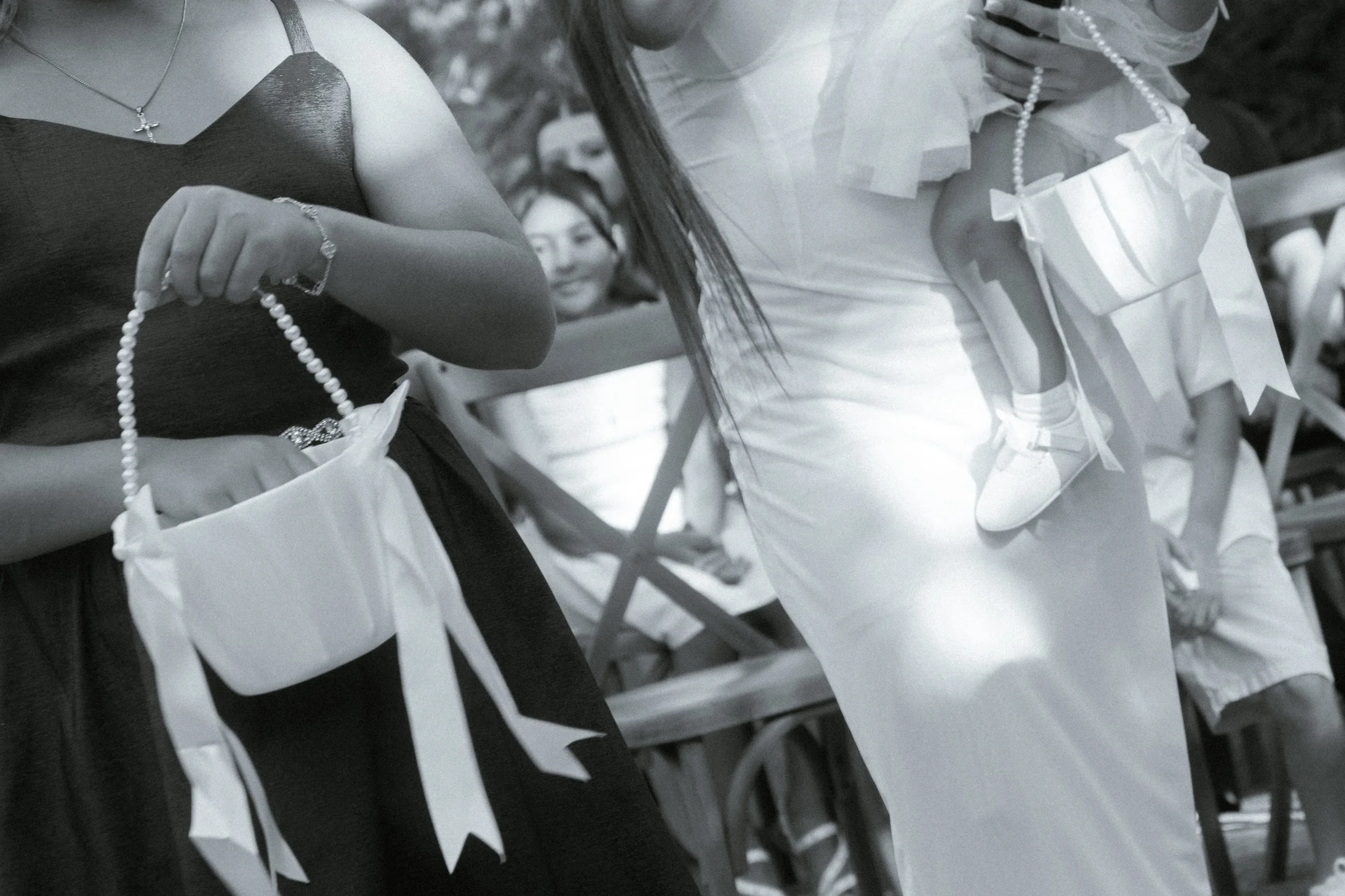 A woman in a dark dress holding a string of pearls, with another person wearing a white dress with large bows and pearl necklace, at an outdoor event, with chairs in the background.