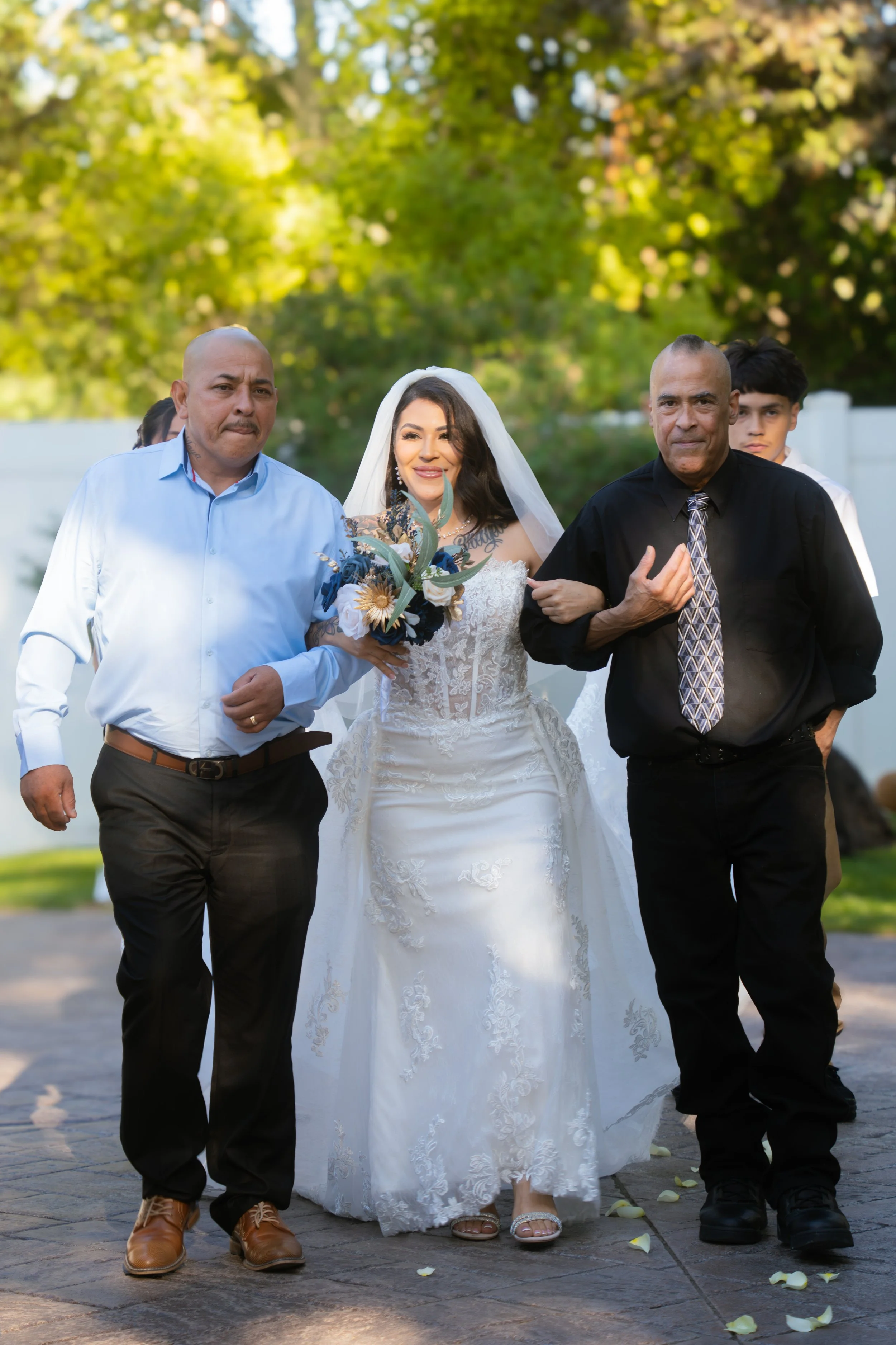 A bride in a white wedding dress with a veil walking with two men, possibly her father and a friend, during a wedding ceremony outdoors with greenery in the background.