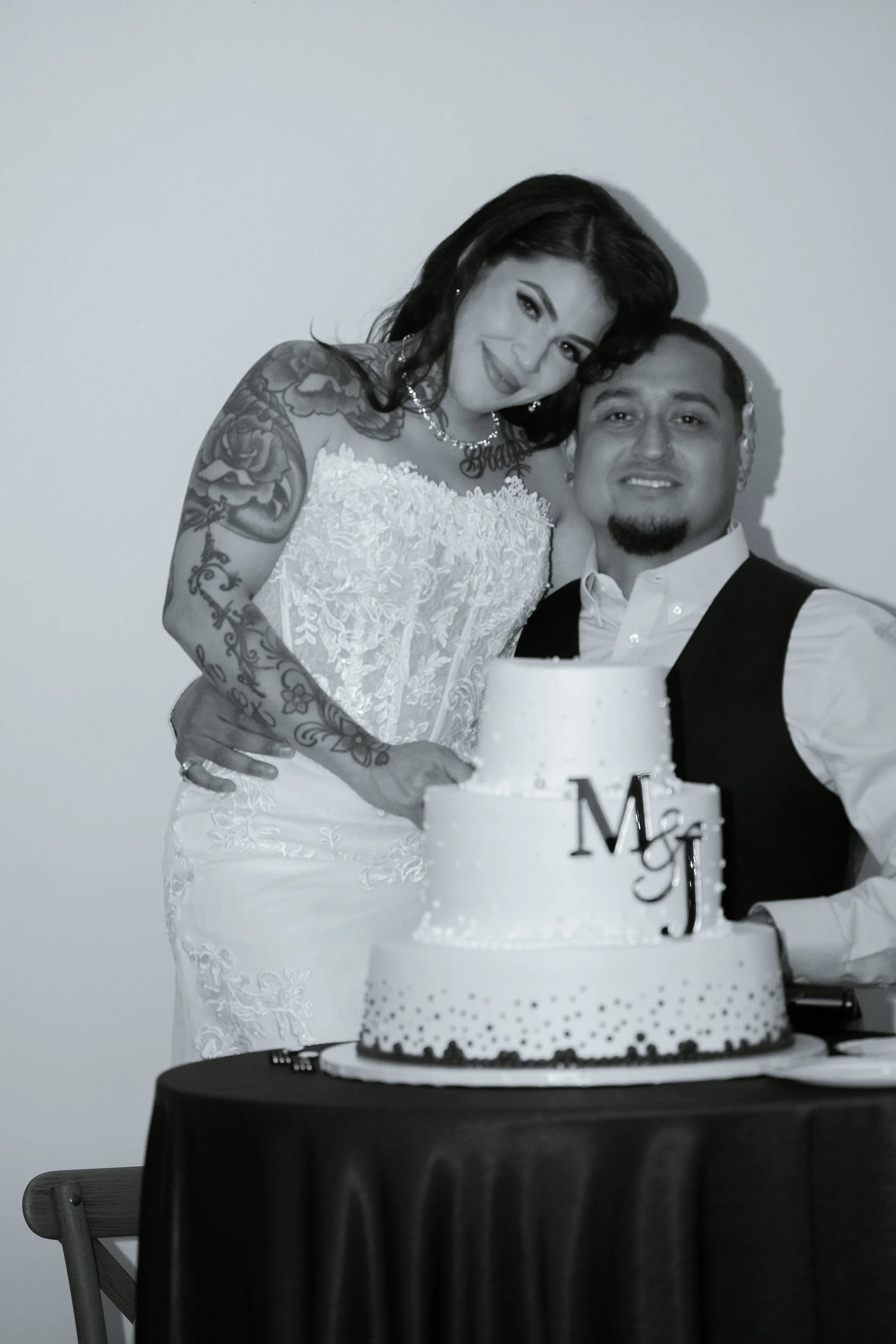 A bride and groom smiling at a wedding celebration, standing behind a decorated wedding cake with a "Mr & Mrs" topper, black tablecloth, and a plain wall background.