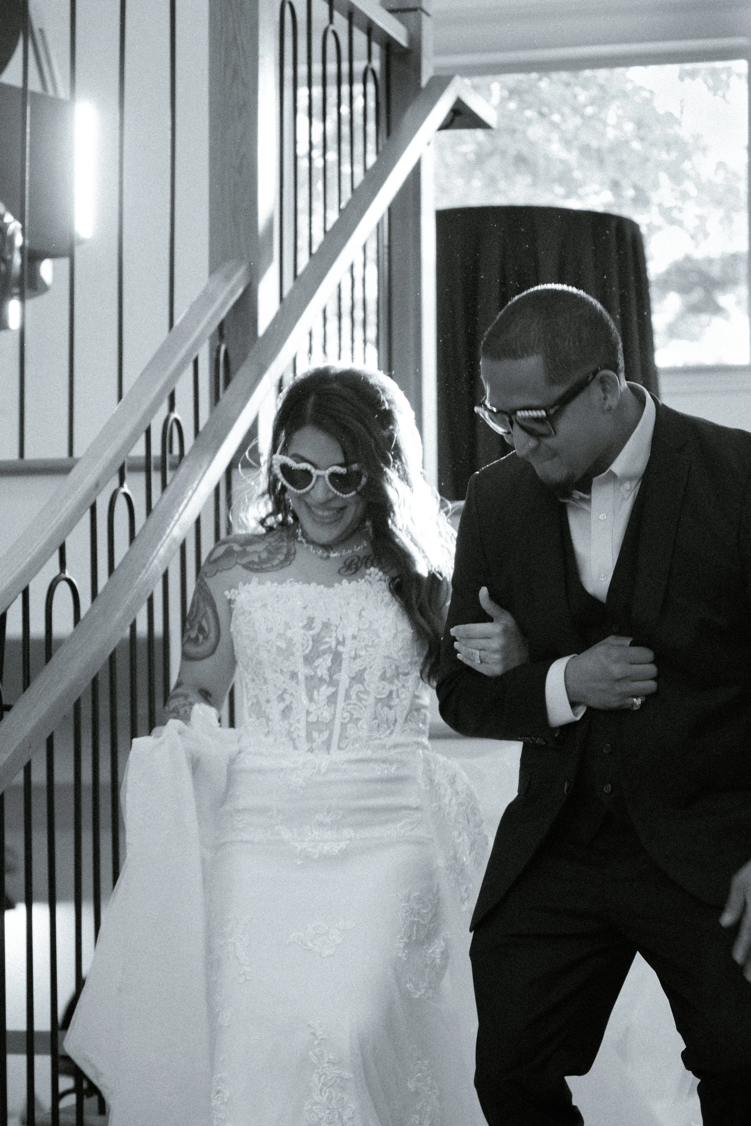 A black and white photo of a bride and groom at their wedding ceremony. The bride is wearing a lace wedding gown and heart-shaped sunglasses, smiling while looking down. The groom is in a tuxedo with a bow tie and glasses, looking at the bride while 