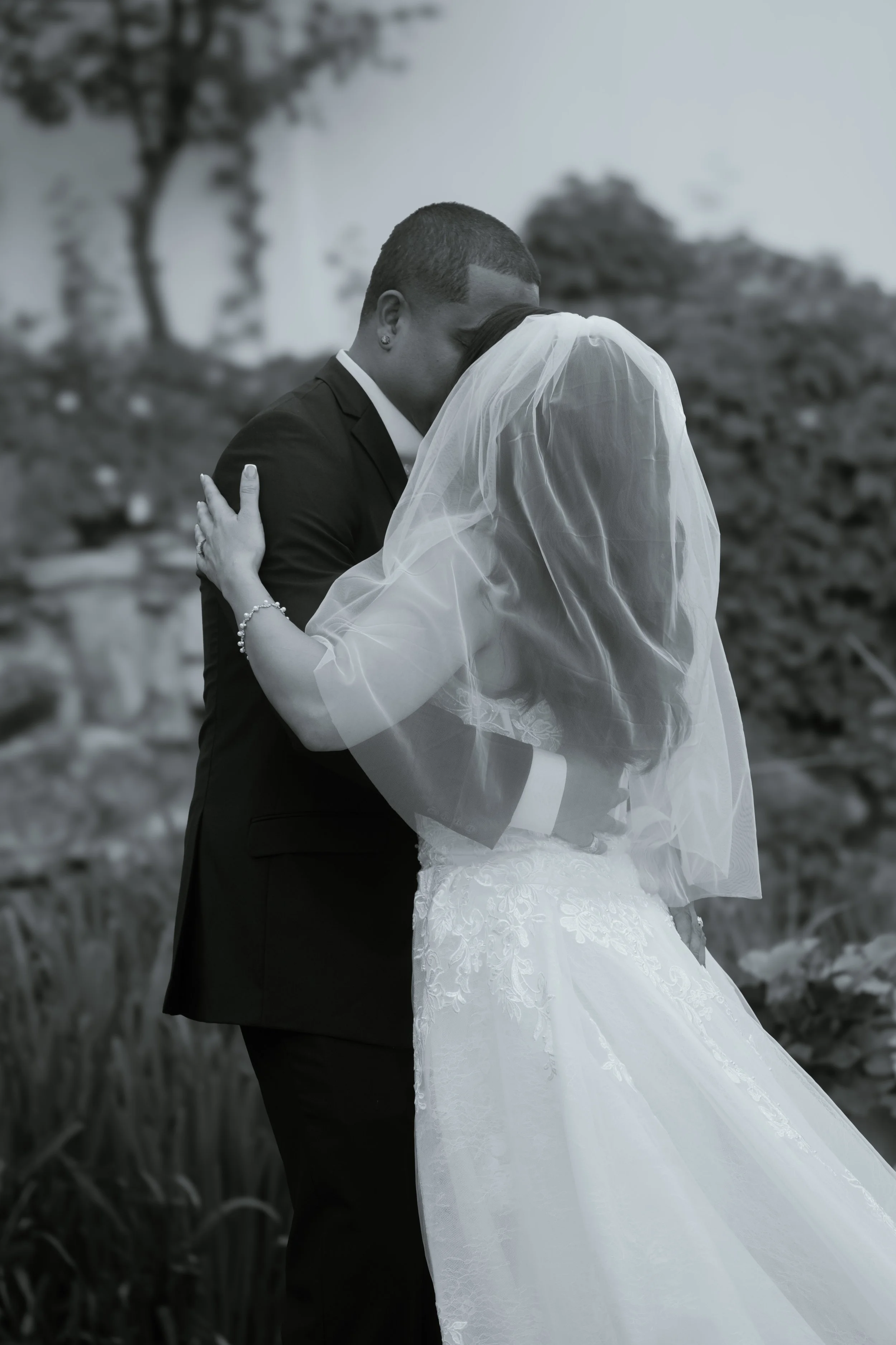 A bride and groom sharing an intimate kiss outdoors, with trees and plants in the background.
