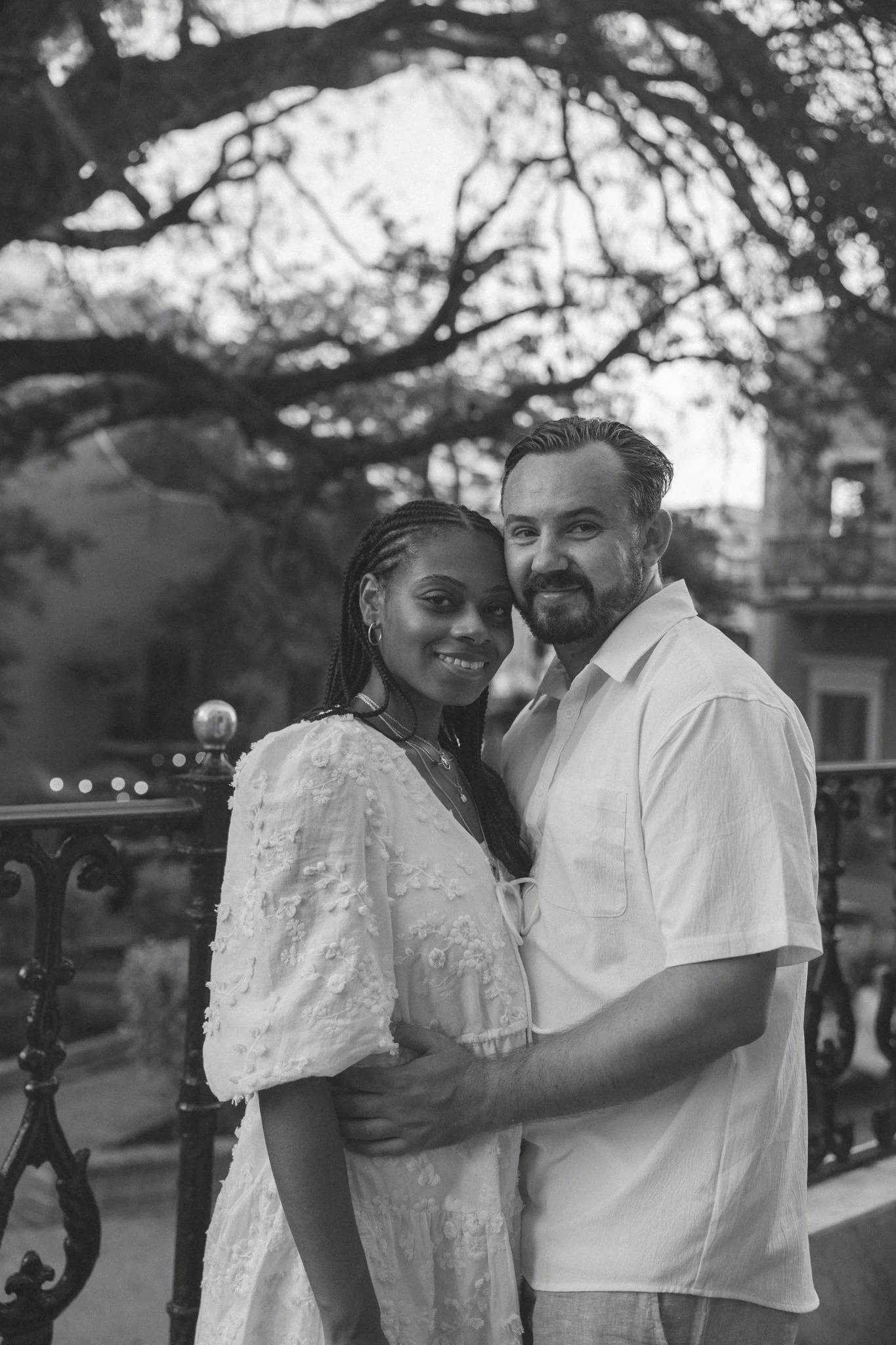 A black-and-white photo of a smiling couple standing close together outdoors, with a large tree and a fence in the background.