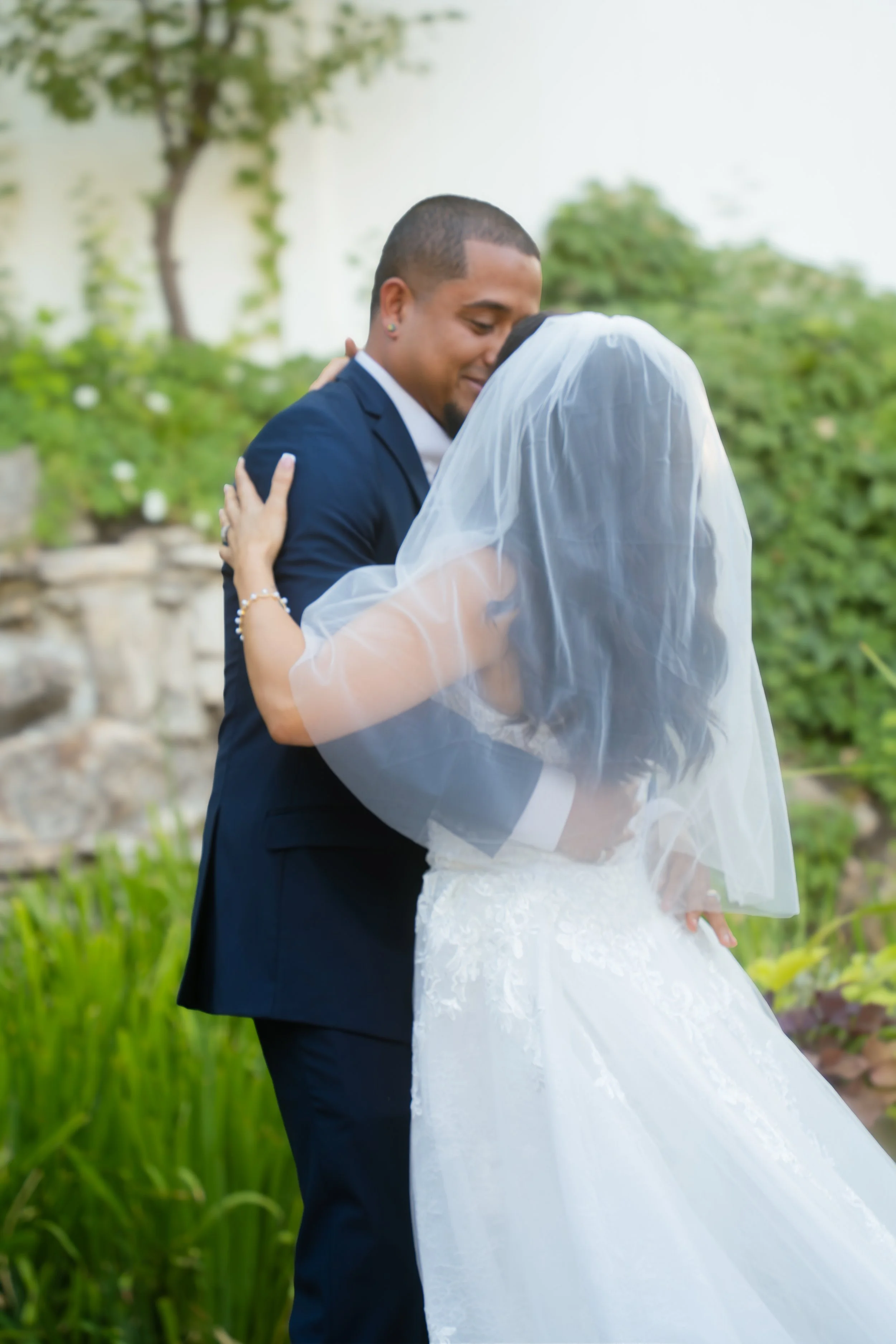 A couple in wedding attire embracing outdoors, with the woman in a white lace wedding dress and veil, and the man in a blue suit, in a lush garden setting.