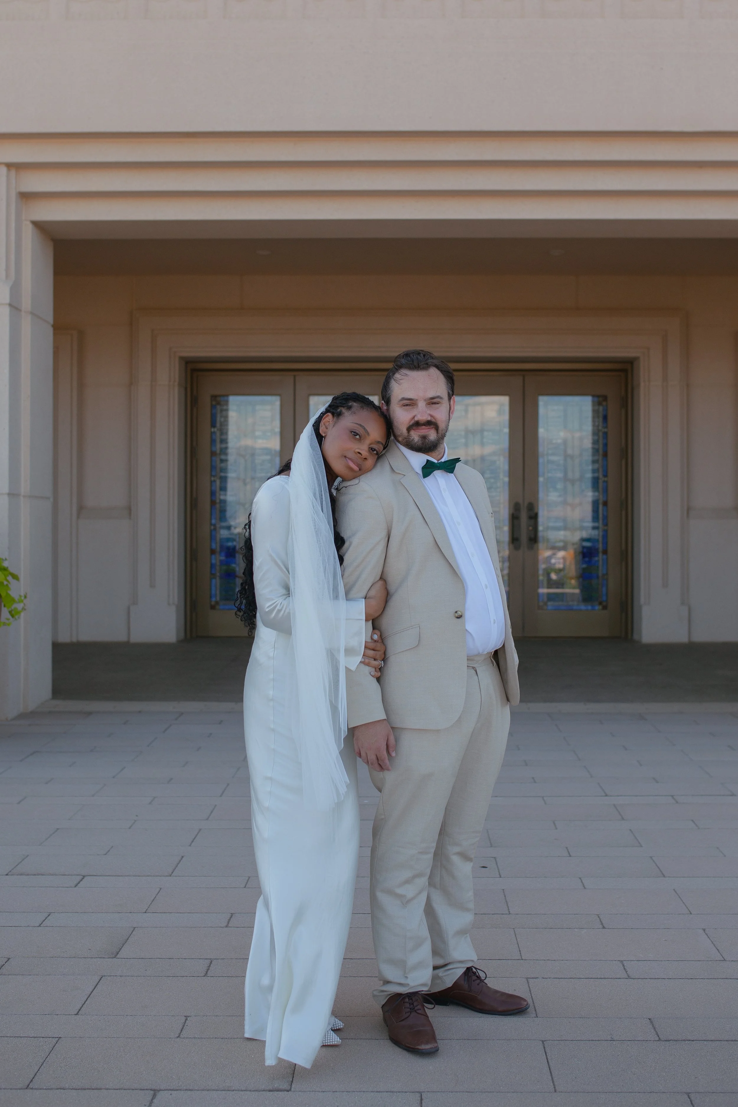 A Black woman in a white suit with a veil leaning her head on a White man in a beige suit with a green bow tie, standing in front of a beige building with glass doors.