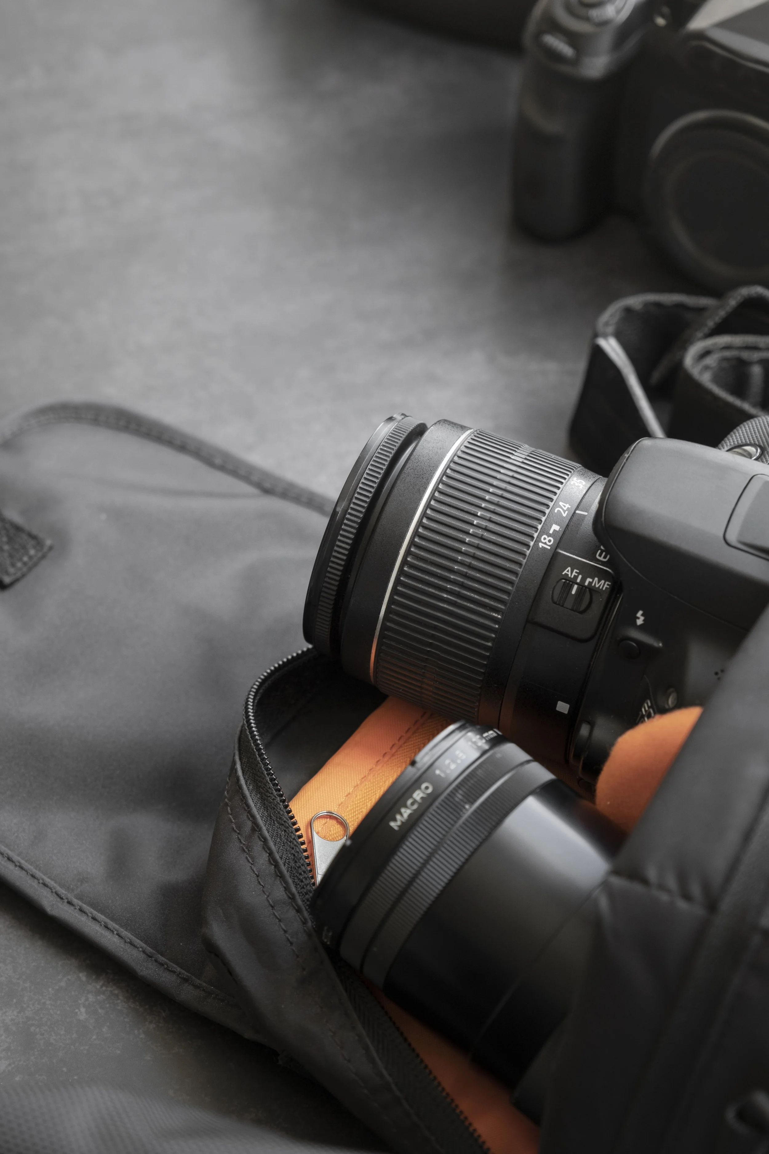 Close-up of a camera with a zoom lens inside a black camera bag, along with an orange item and a silver key. The camera appears to be a mirrorless or DSLR type.