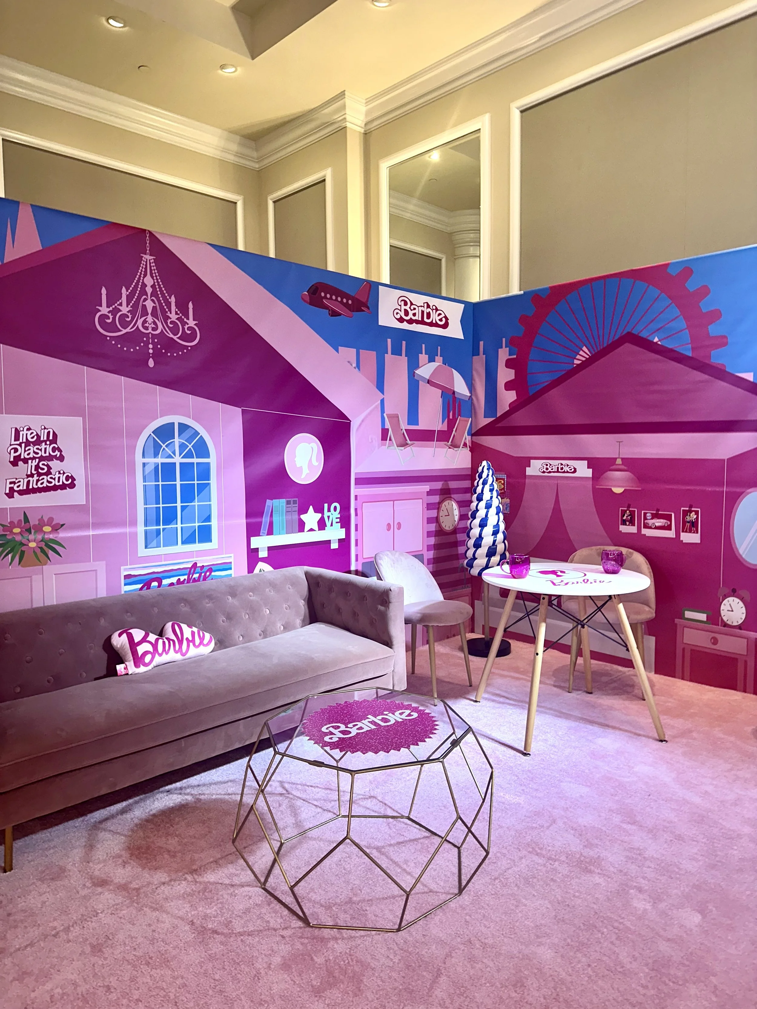Colorful Barbie-themed room with pink and purple decor, featuring a gray velvet sofa, a round white table, and wall murals with Barbie graphics and cityscape images.