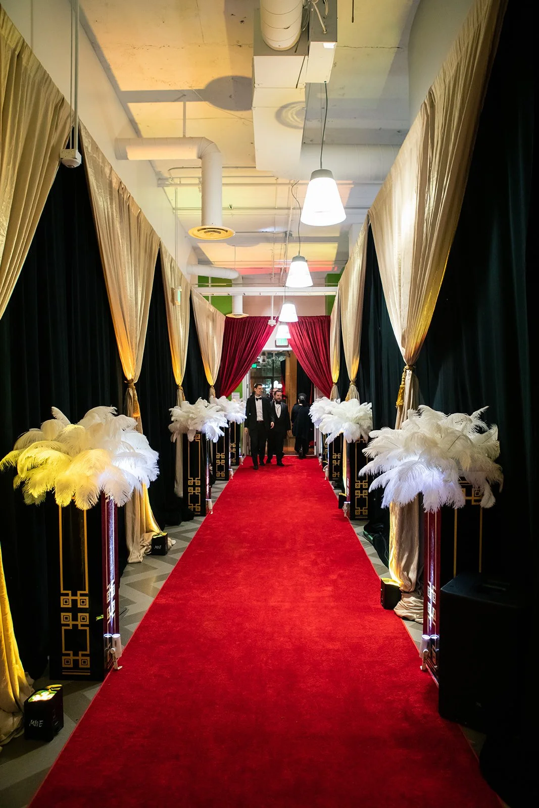 Elegant hallway with a red carpet, gold and black curtains, and white feather decorations on stands leading to a doorway with red curtains and guests in formal wear