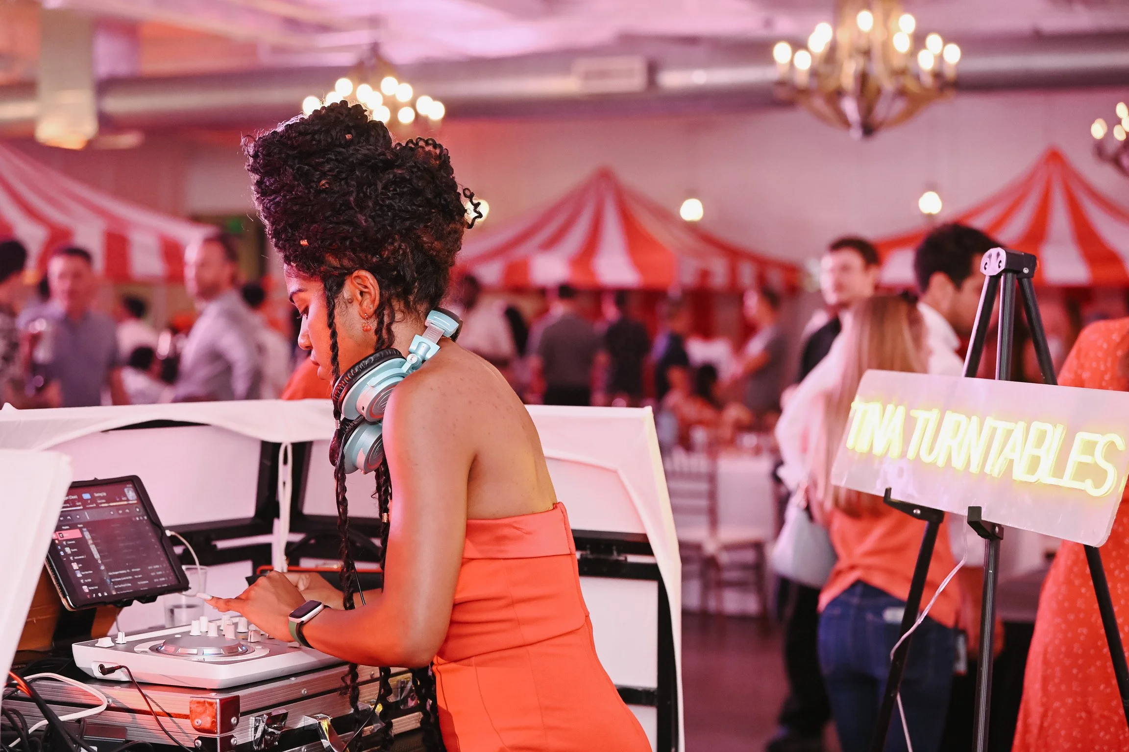 A female DJ with curly hair and wearing a strapless orange dress is mixing music at a lively event with red and white striped tents and guests in the background.