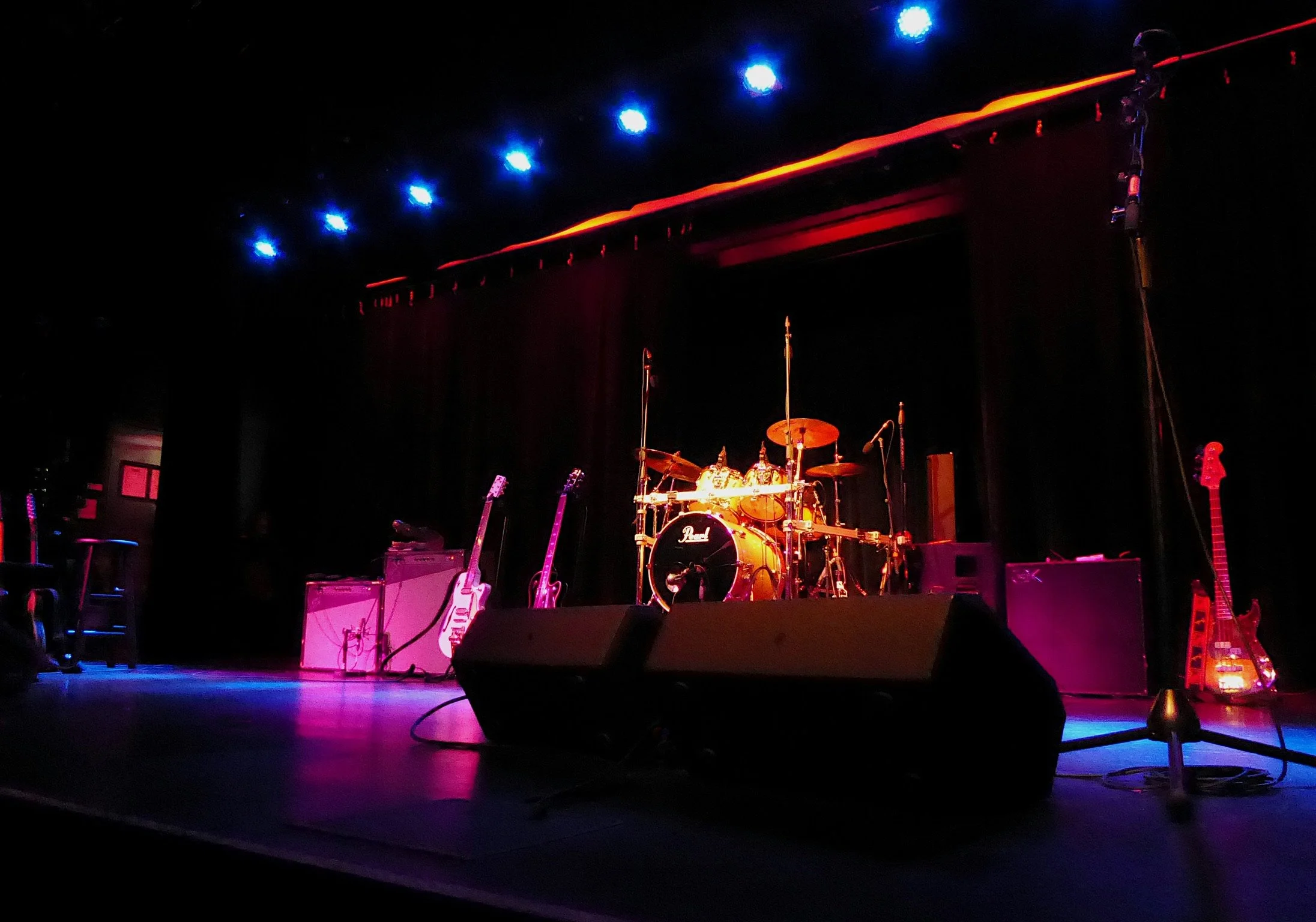 Empty stage with musical instruments including a drum set, guitars, and amplifiers, illuminated by blue and pink stage lights.