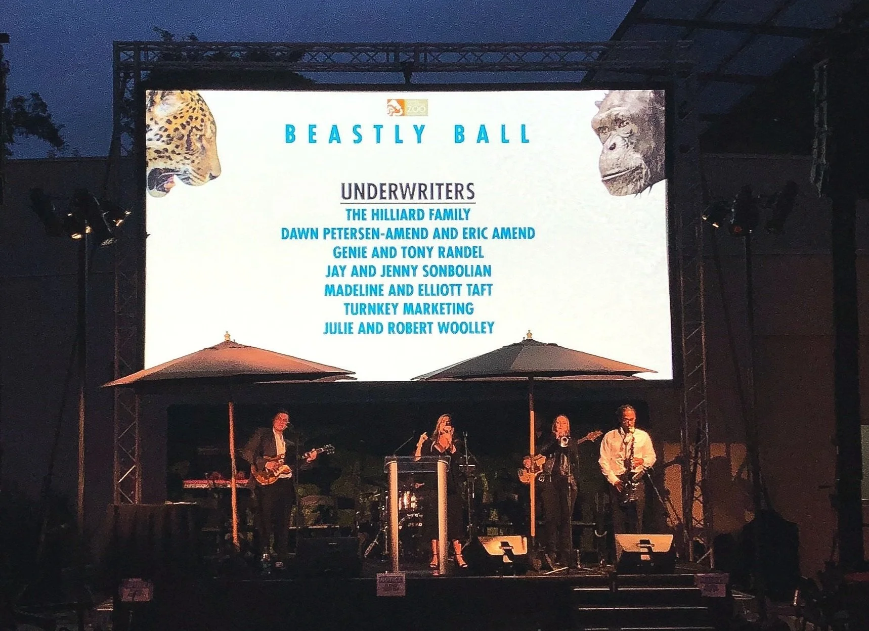 Stage with band performing and a large screen displaying the title 'Beastly Ball' and a list of underwriters at the zoo, with images of a leopard and a lion on the top corners.
