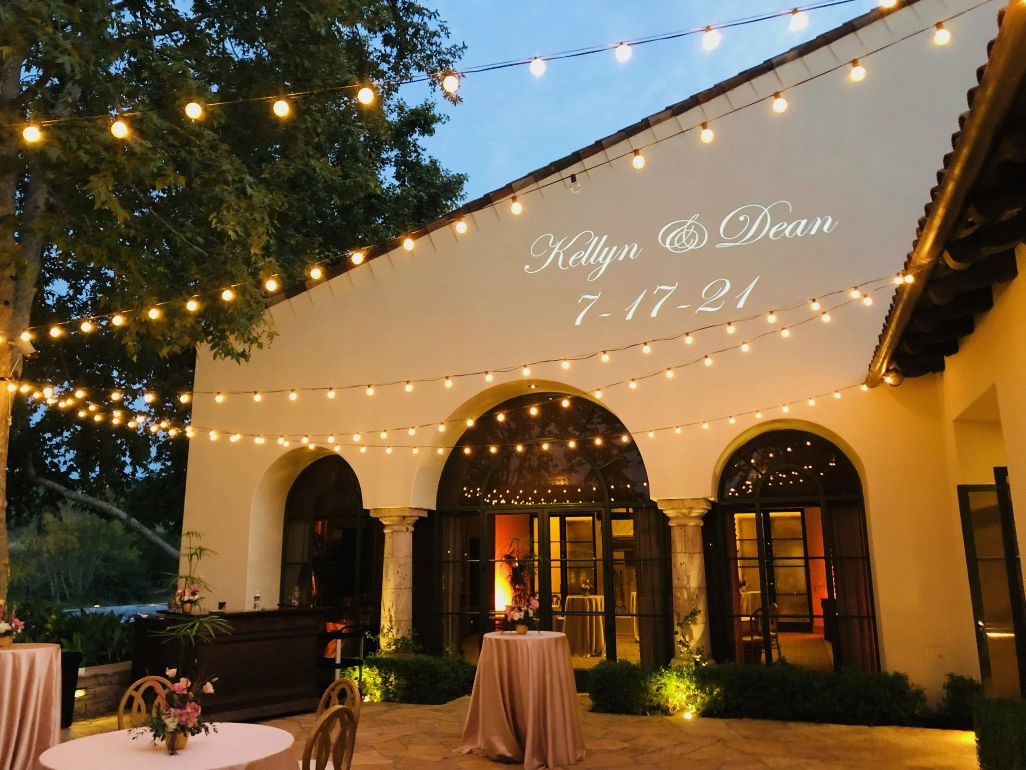 Outdoor wedding reception with string lights, floral arrangements on tables, and a building with projected text reading "Kellyn & Dean 7.17.21".