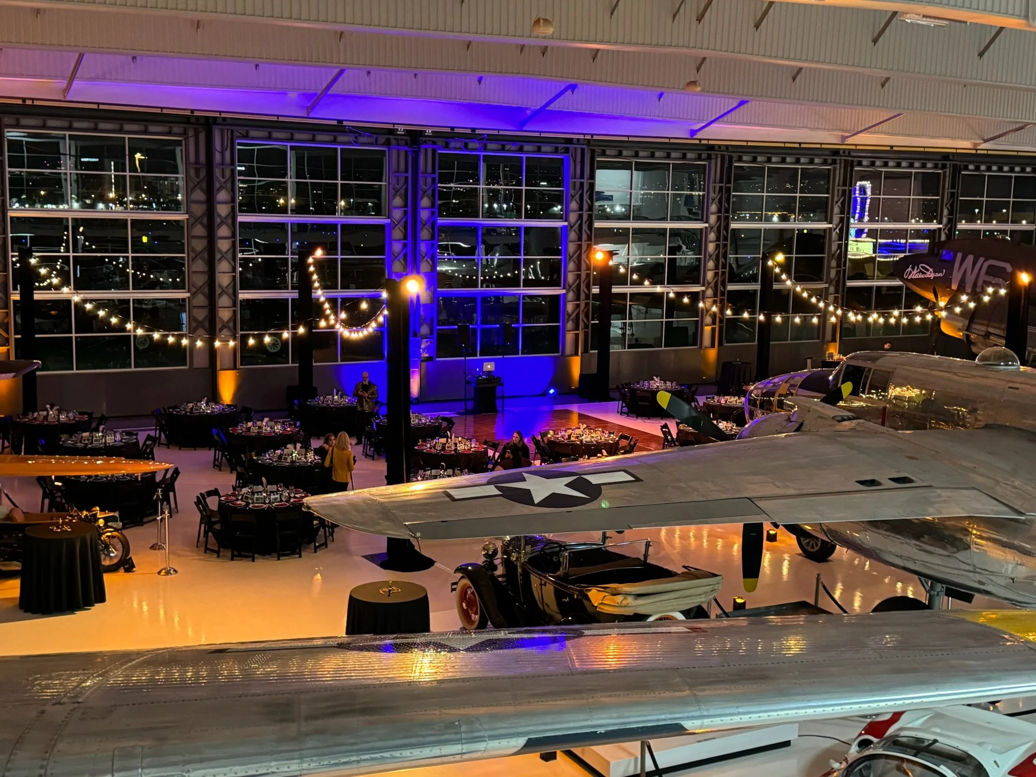 Interior of an aviation-themed event space with vintage aircraft on display, decorated with string lights and banquet tables set for a dinner event.