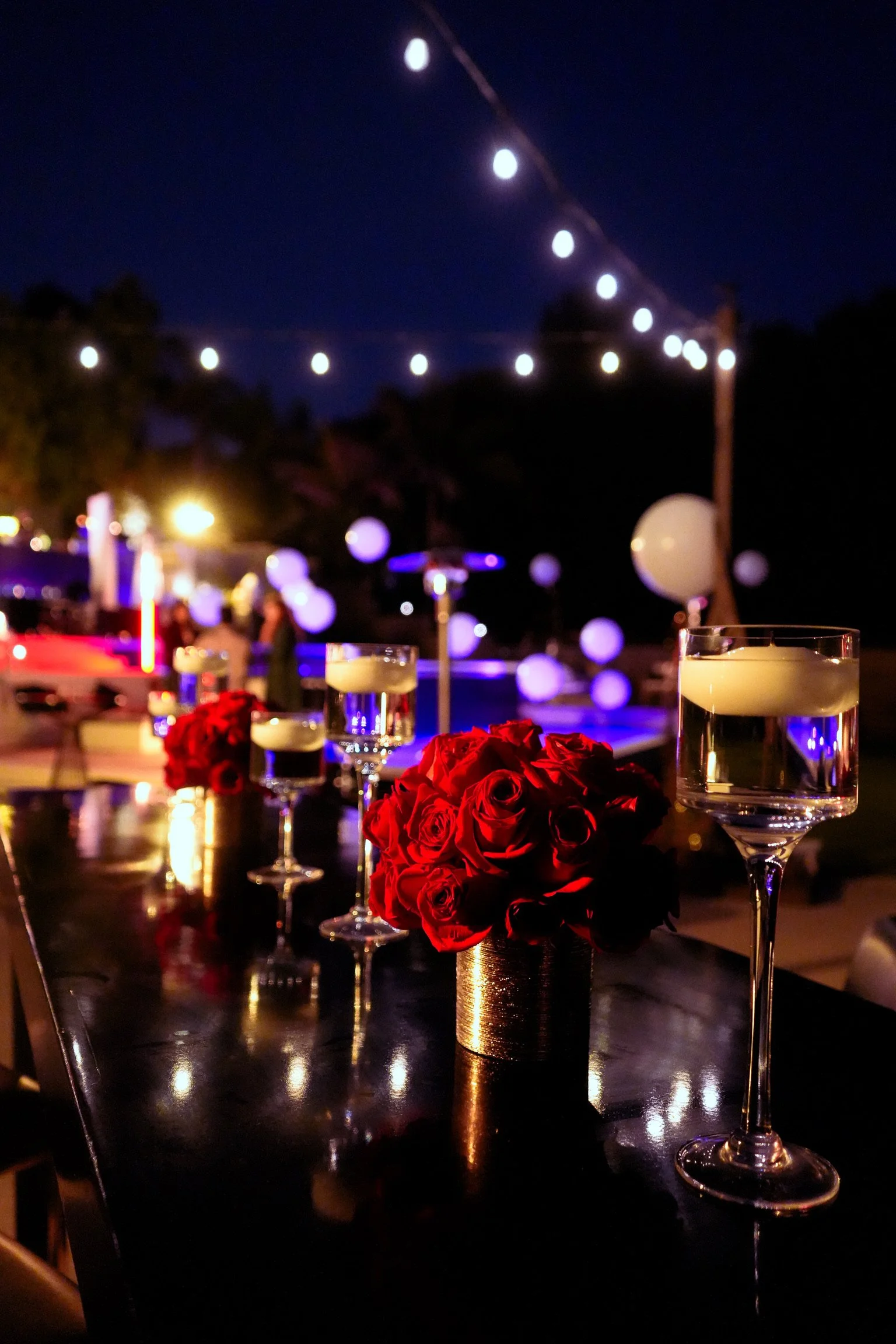 An evening outdoor party scene with string lights, white balloons, red roses in a gold vase, and glasses of water on a reflective black table.