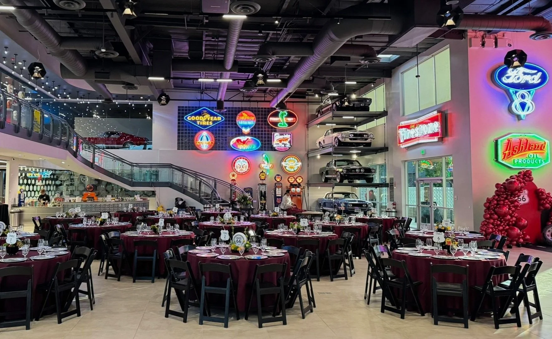 Event space decorated with vintage neon signs and classic cars on display, with round tables covered in burgundy tablecloths and black chairs, set for a meal.
