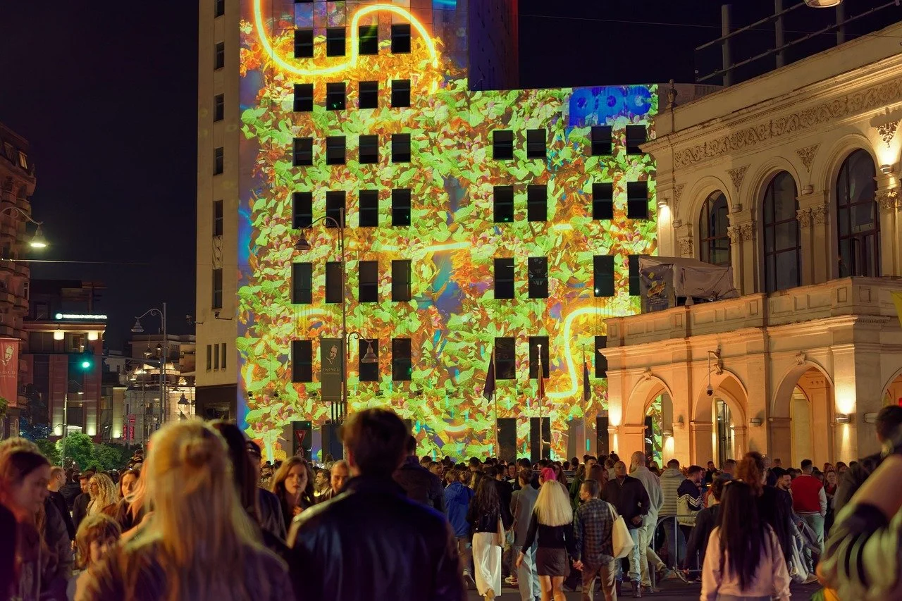 Nighttime city scene with a large crowd gathered in front of a building displaying colorful, animated projections on its facade in the background. The projections include vibrant patterns and shapes, with a notable bright yellow outline in the upper part of the building. People are standing and walking, enjoying the visual display.