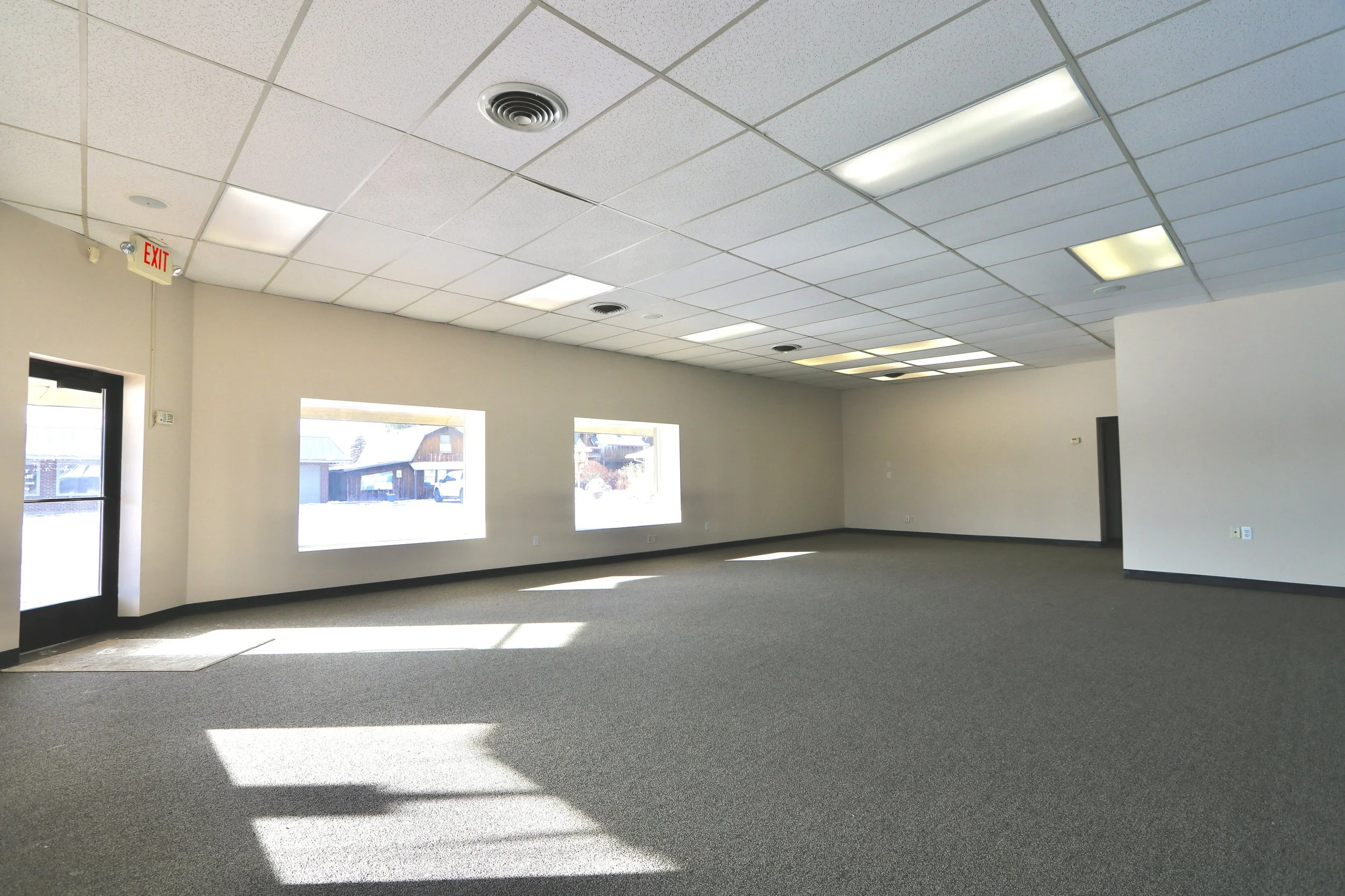 Empty commercial space with beige walls, gray carpet flooring, multiple ceiling lights, and large rectangular windows letting in sunlight.